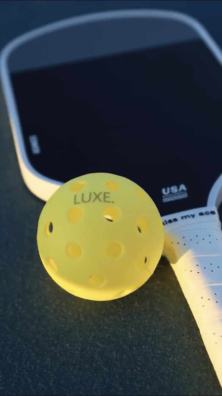 A close-up of the LUXE Pickleball Dinker, a USA Pickleball Approved carbon fiber paddle in black and white, lies on a dark court with a bright yellow LUXE pickleball resting against its overgrip-wrapped white grip.