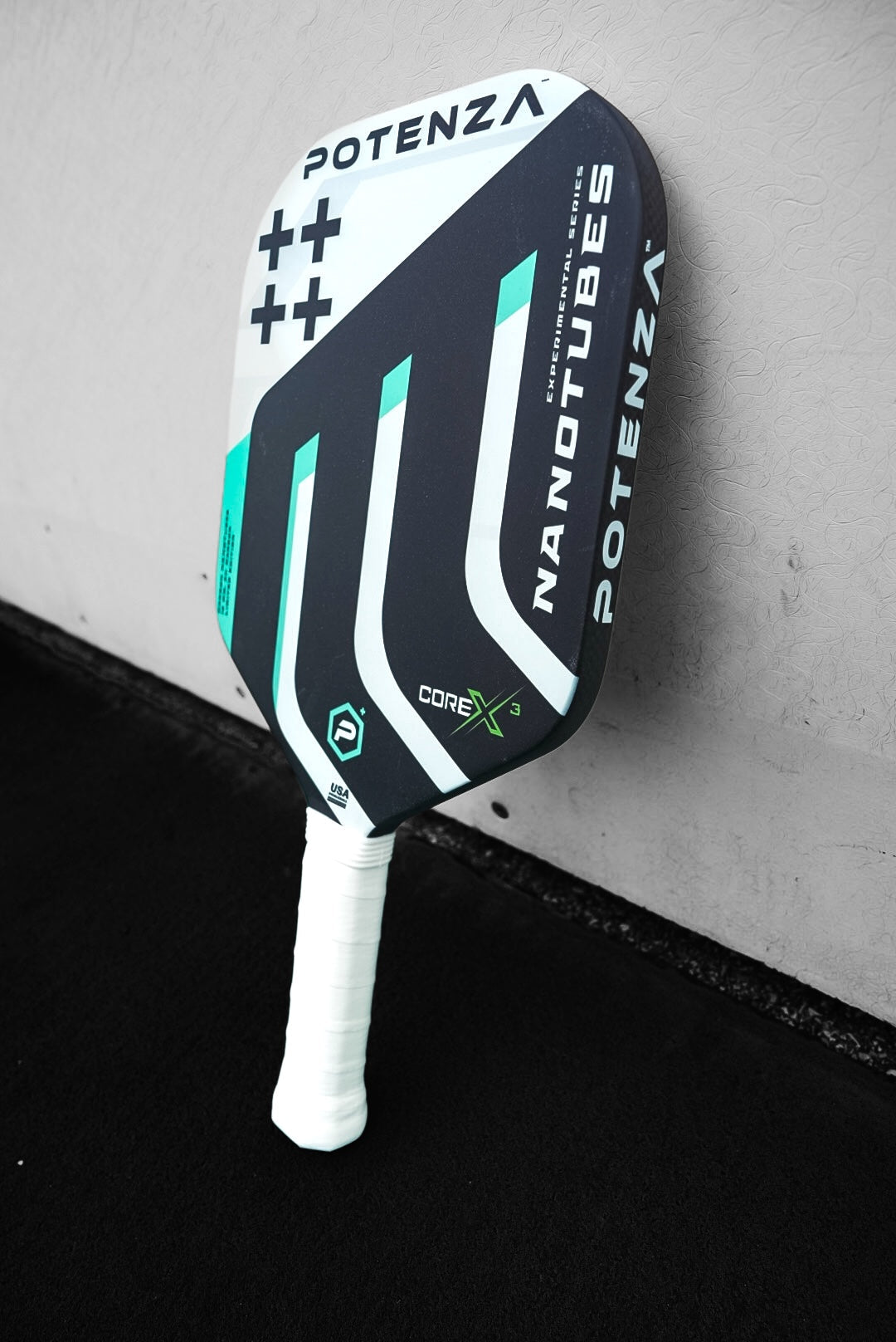 The Potenza Pickleball Carbon Nanotubes+ COREx3 paddle stands upright with a black, white, and teal geometric design, durable carbon nanotube construction, a white grip handle, and bold branding against a gray wall.