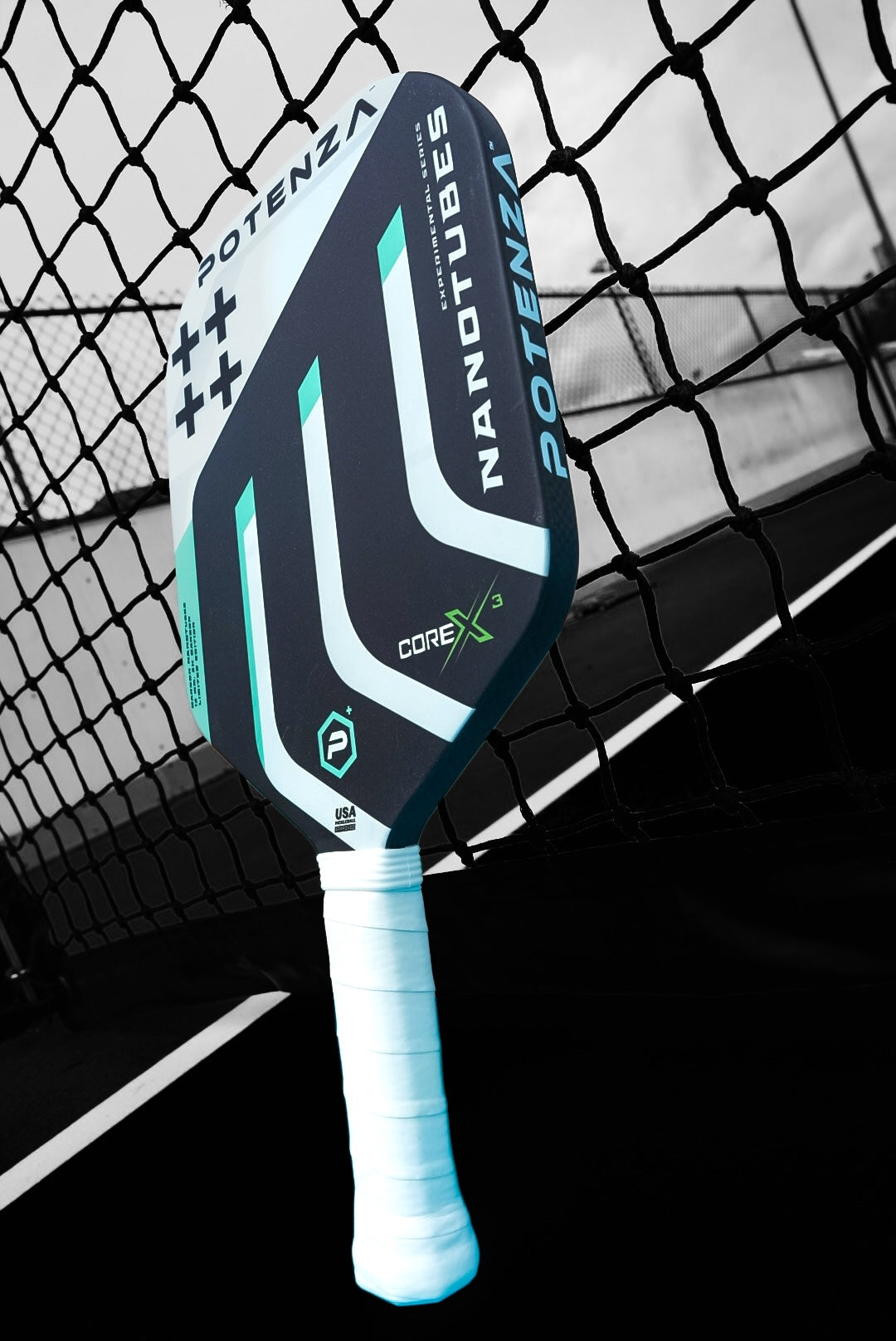 A close-up shows the Potenza Pickleball Carbon Nanotubes+ COREx3 paddle in black and teal, with white grip tape and geometric designs, leaning against a net on an outdoor court with visible lines and fencing in the blurred background.