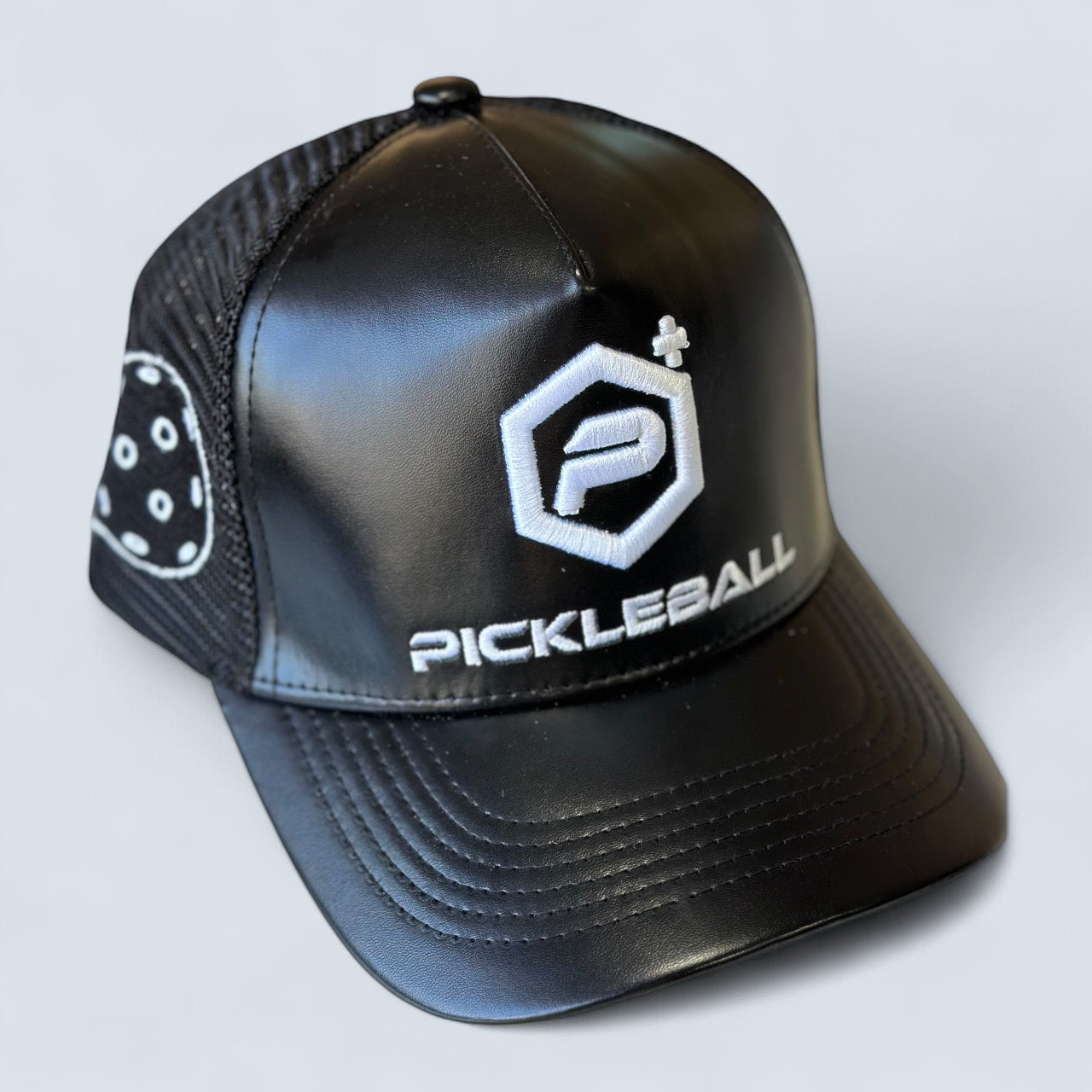 The Potenza Pickleball Leather Logo Trucker Hat features mesh sides, a shiny black front panel, and a white embroidered stylized “P” with “Pickleball+”, plus a sporty pickleball image on the side for the ultimate pickleball hat look.