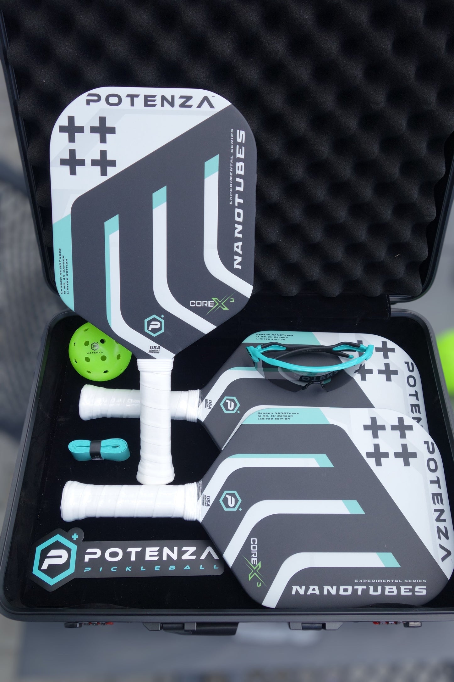 An open black case displays three Potenza Pickleball Carbon Nanotubes+ COREx3 paddles with geometric designs in white, black, teal, and grey, a yellow wiffle ball, teal grip tape, a teal wristband, and teal-and-black sports sunglasses.