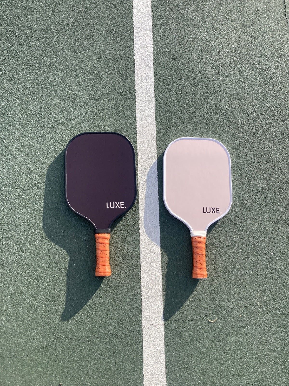 Two Aesthetic Solids - Ace paddles from LUXE Pickleball rest on a green court split by a white line. Both have wooden handles and are Pickleball Approved, with one in black and the other in white near the base.