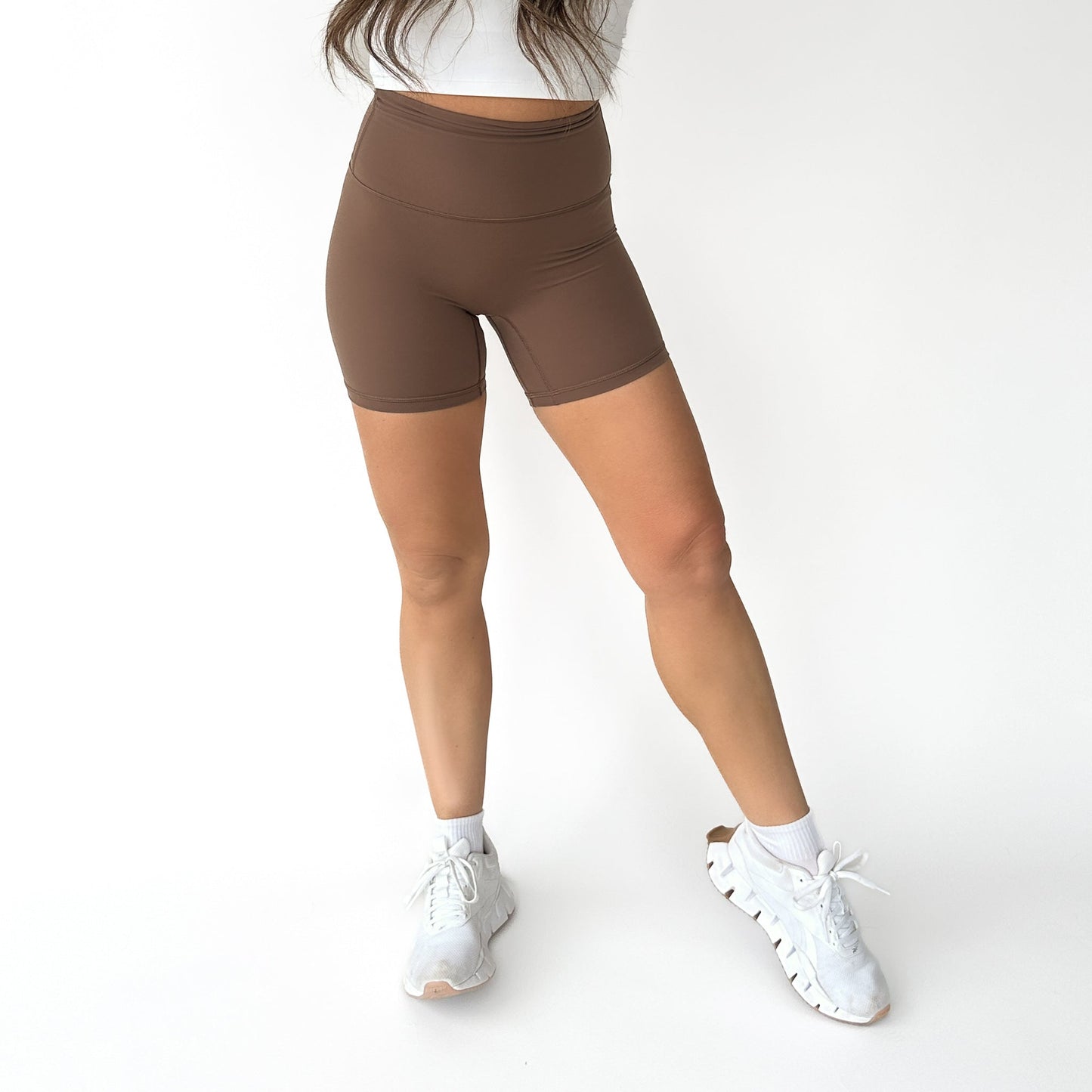 A woman stands against a white background wearing DYM Athletics AIRE Ultra-Soft High Waist Biker Shorts | 5", paired with a white cropped top, ankle socks, and sneakers. She is partially cropped from the waist up, arms relaxed and one leg bent.