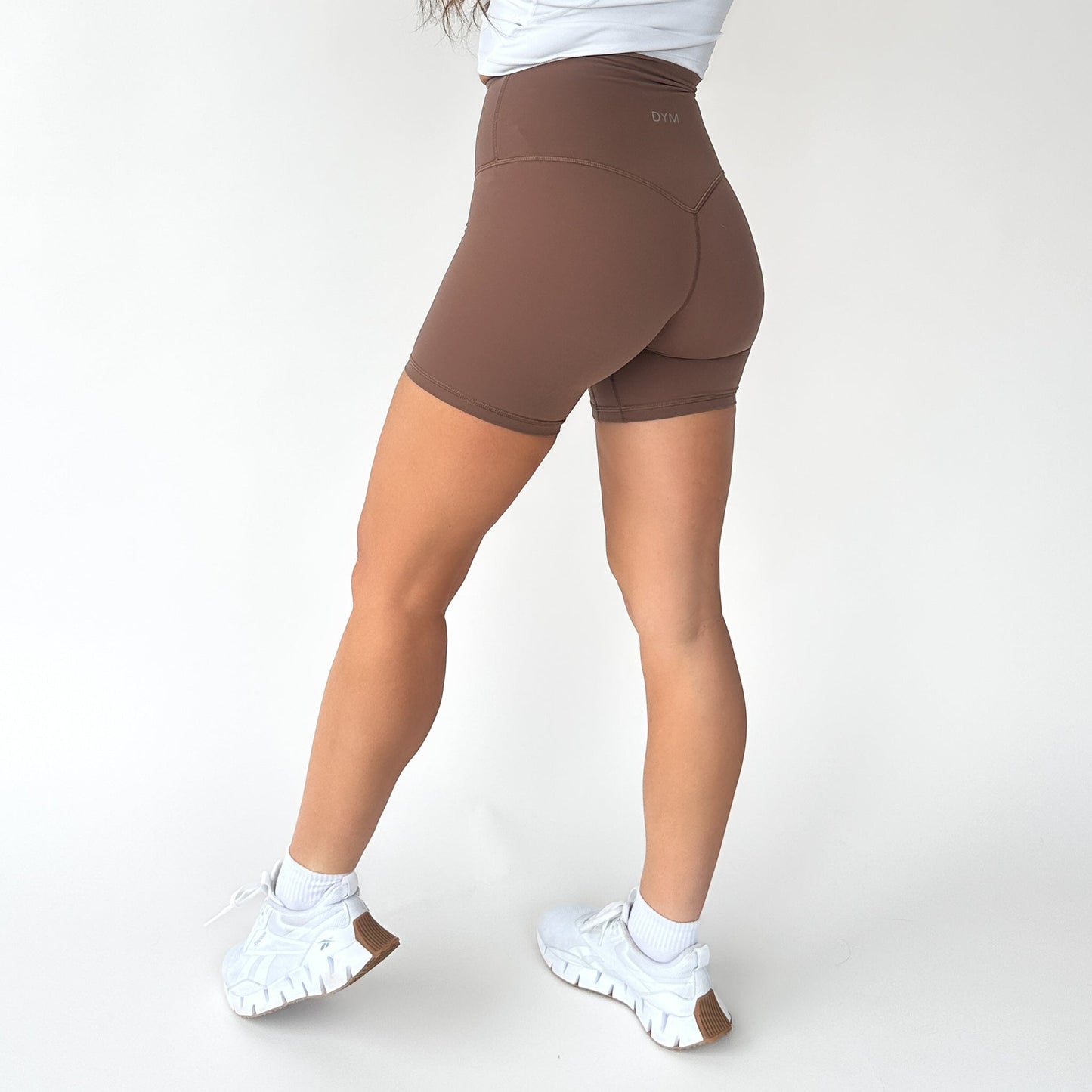 A person stands on a white background, showcasing their lower body in DYM Athletics AIRE Ultra-Soft High Waist Biker Shorts | 5", paired with white socks and sneakers.