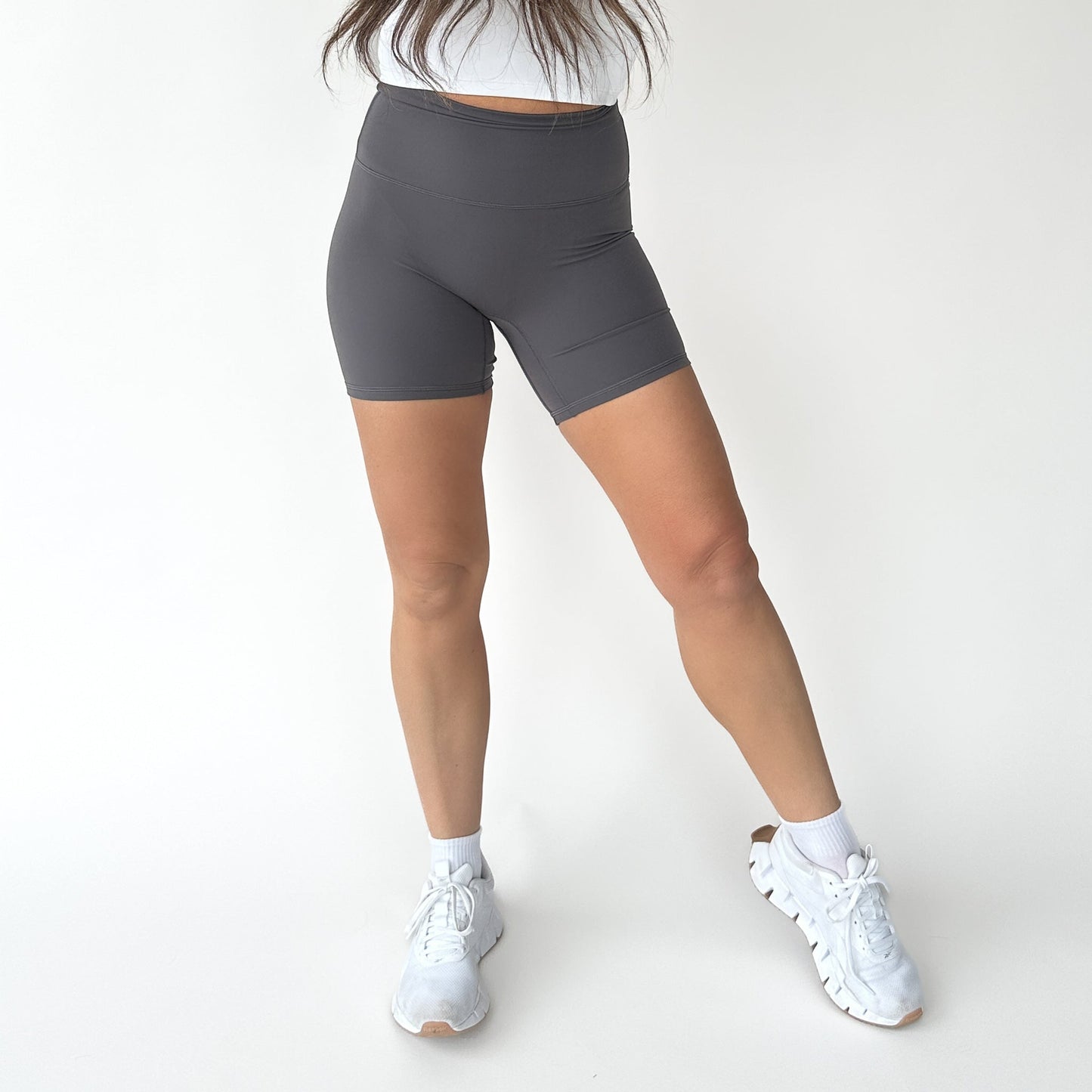 A person stands against a plain background wearing DYM Athletics AIRE Ultra-Soft High Waist Biker Shorts | 5" styled with a white top, white crew socks, and white athletic sneakers. Their head isn’t visible; legs are slightly apart with one knee bent.