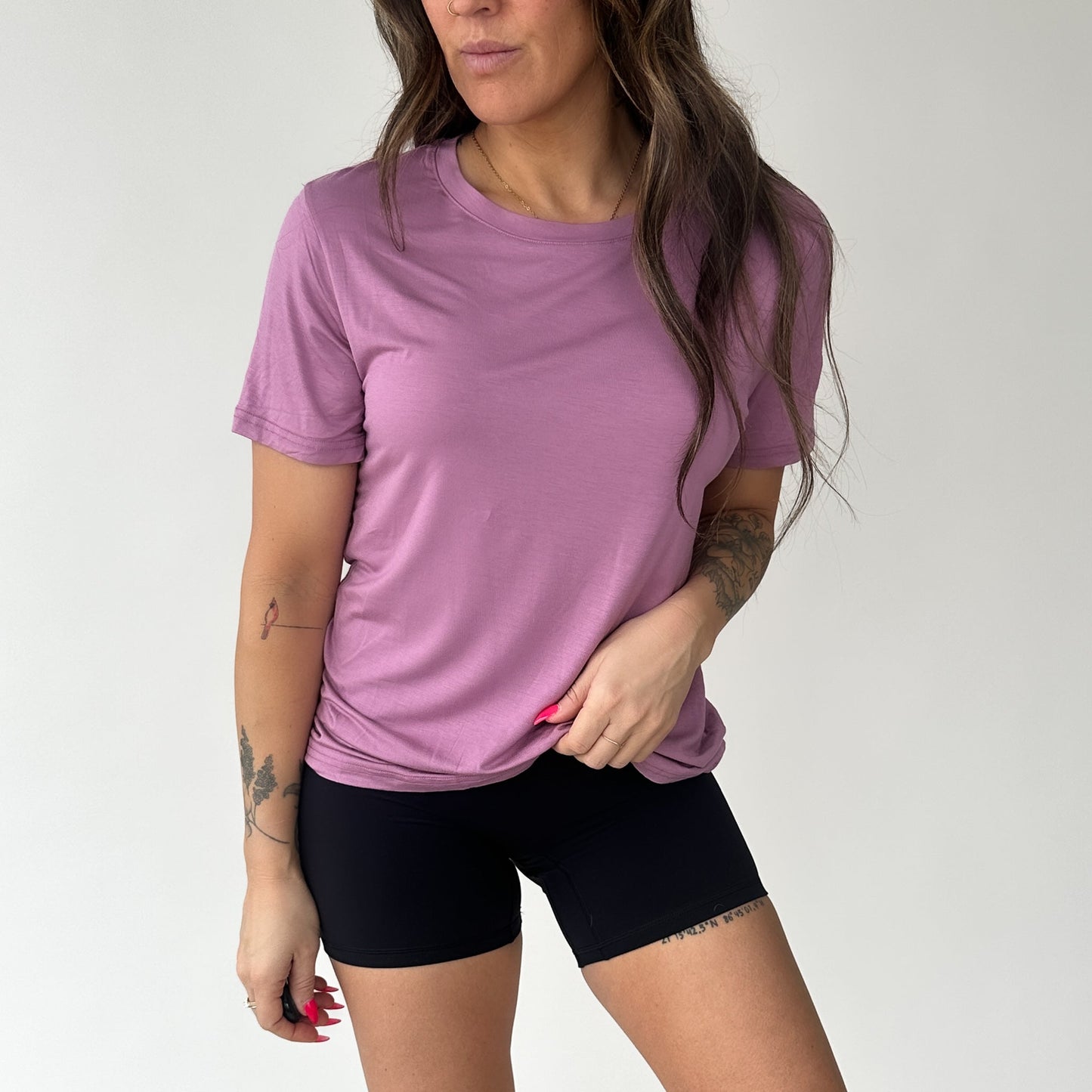 A person with long wavy hair wears the DYM Athletics Breathe Bamboo T-Shirt in mauve, paired with black fitted shorts. Visible tattoos and long pink nails add detail as they pose against a plain white background, one hand on their hip.