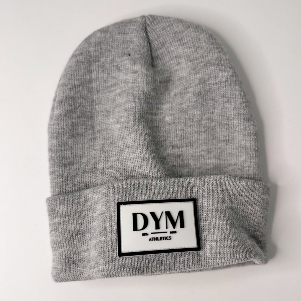 The DYM Beanie by DYM Athletics is a light gray, ribbed knit hat with a cozy folded brim and a white patch labeled DYM ATHLETICS in bold black letters, shown on a white background. Perfect for cool weather. Now on SALE.