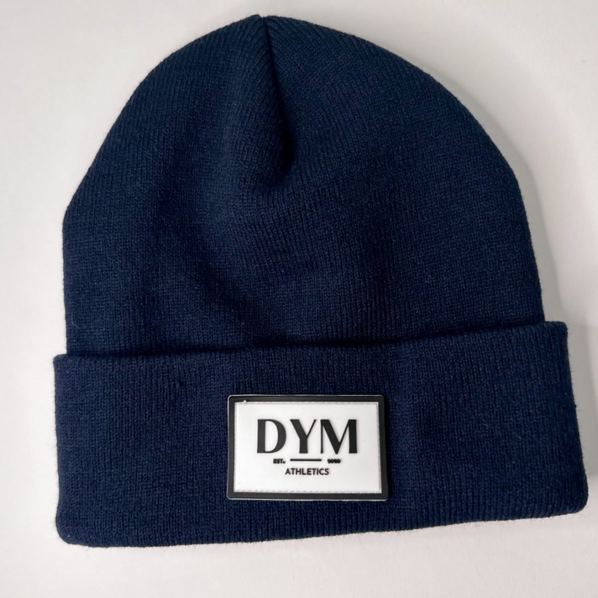 The DYM Beanie | SALE from DYM Athletics is a cozy navy knit hat with a folded brim and a retro rectangular white-and-black patch reading “DYM ATHLETICS.” Perfect for cool days, shown here against a white background.
