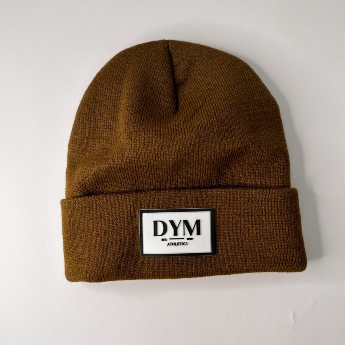 The DYM Beanie by DYM Athletics is shown on a white background. This cozy brown knit beanie features a folded brim and a retro front patch reading "DYM ATHLETICS," with "ATHLETICS" in a smaller font beneath.