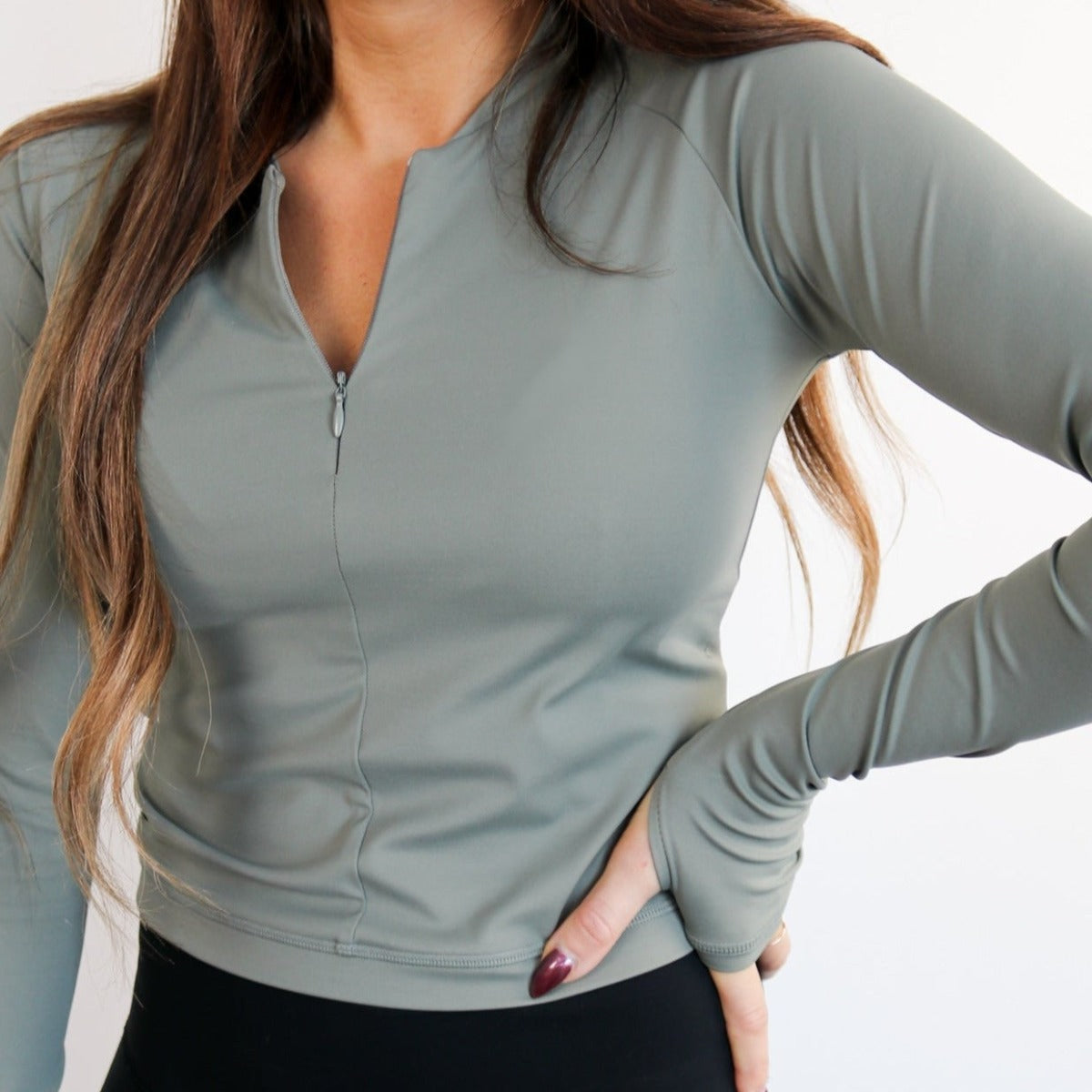 A woman with long brown hair wears the DYM Athletics Elite 1/4 Zip Long Sleeve in light olive green with thumb holes and black high-waisted leggings. The moisture-wicking athletic top is perfect for active days. She stands against a plain light background.