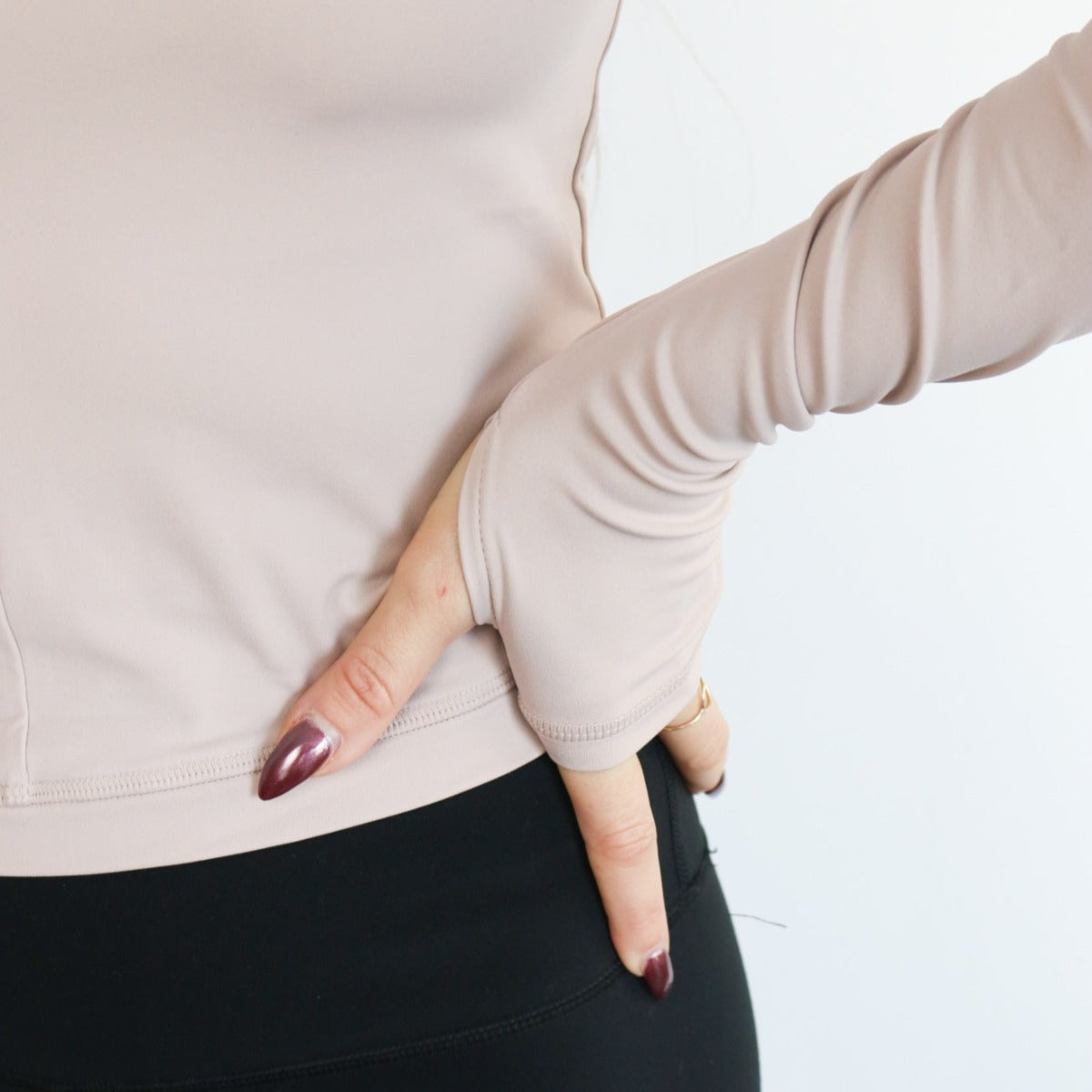 Wearing the DYM Athletics Elite 1/4 Zip Long Sleeve with thumb holes and black pants, a person places their left hand—showing long, dark maroon nails—on their hip. The cropped photo emphasizes the torso and arm on a white background.