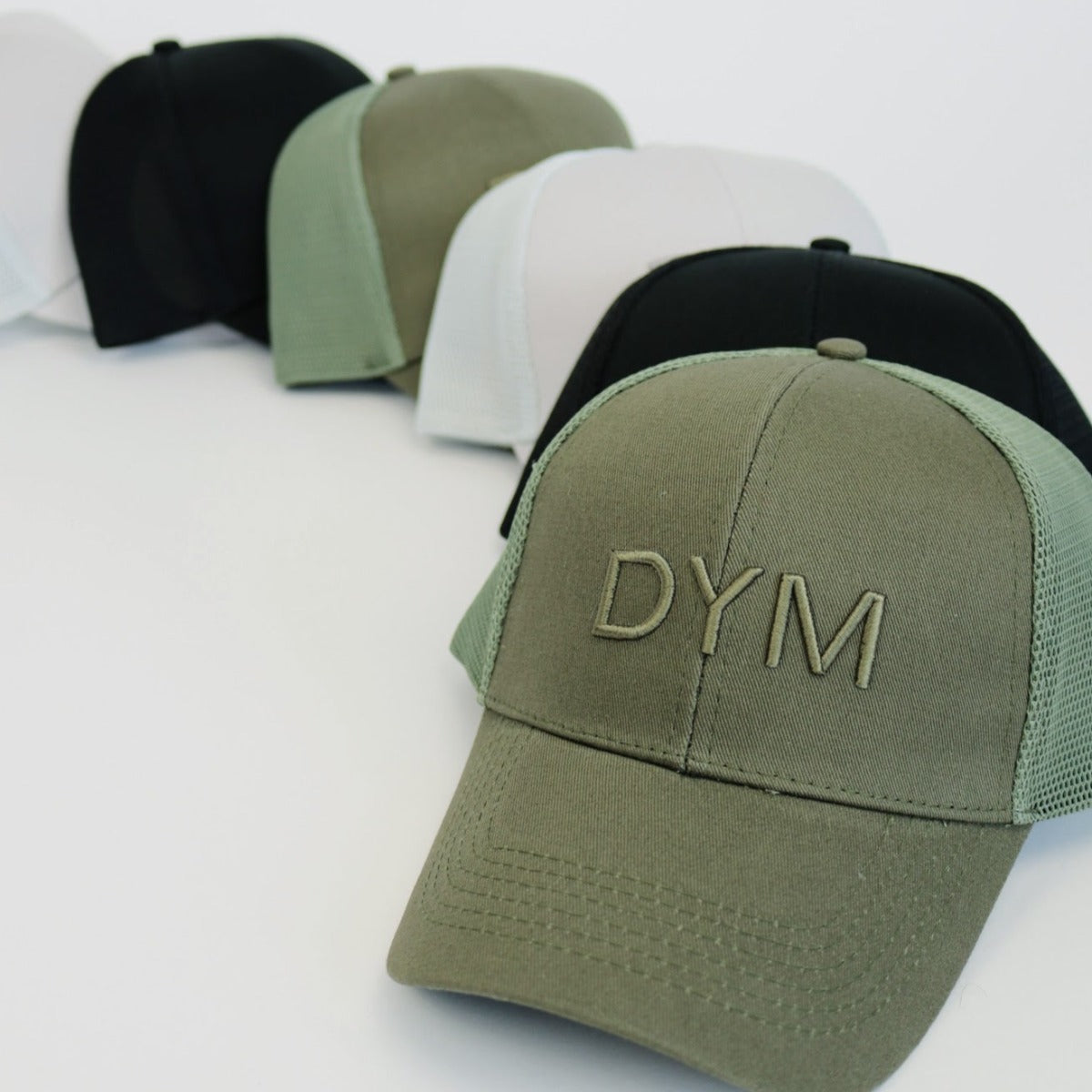 Five DYM Athletics Trucker Hats are arranged diagonally on a white background. The closest is olive green with bold embroidery. All hats have an adjustable snapback and come in black, white, green-and-white, and black-and-white variants.