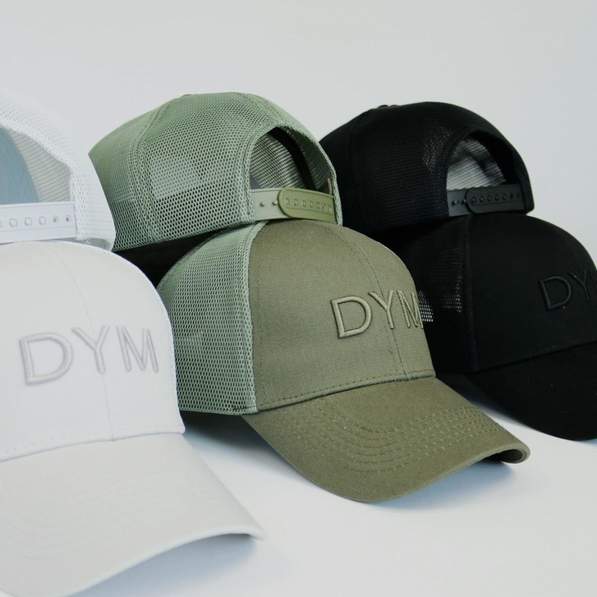 Four DYM Athletics Trucker Hats—white, light green, olive green, and black—are displayed in a row. Each features embroidered "DYM" lettering and an adjustable snapback closure against a plain white background.