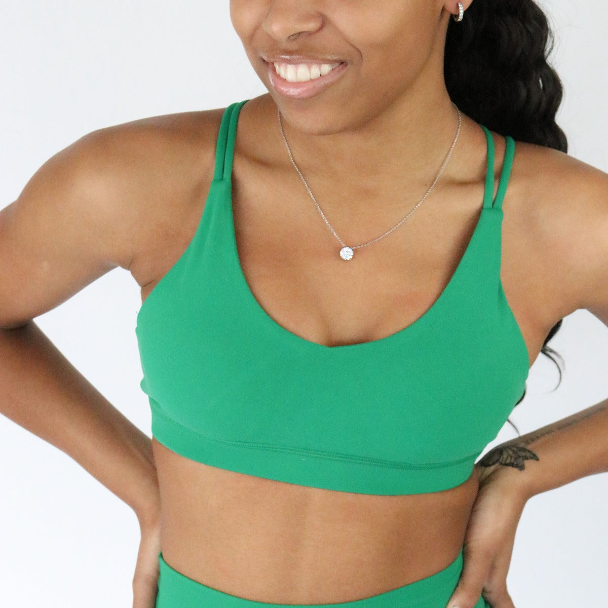 A woman with long dark hair, small hoop earrings, a silver pendant necklace, and a left forearm tattoo smiles in the DYM Athletics AIRE Ultra-Soft Cross Back Sports Bra (A-D cup, medium support) against a plain white background.