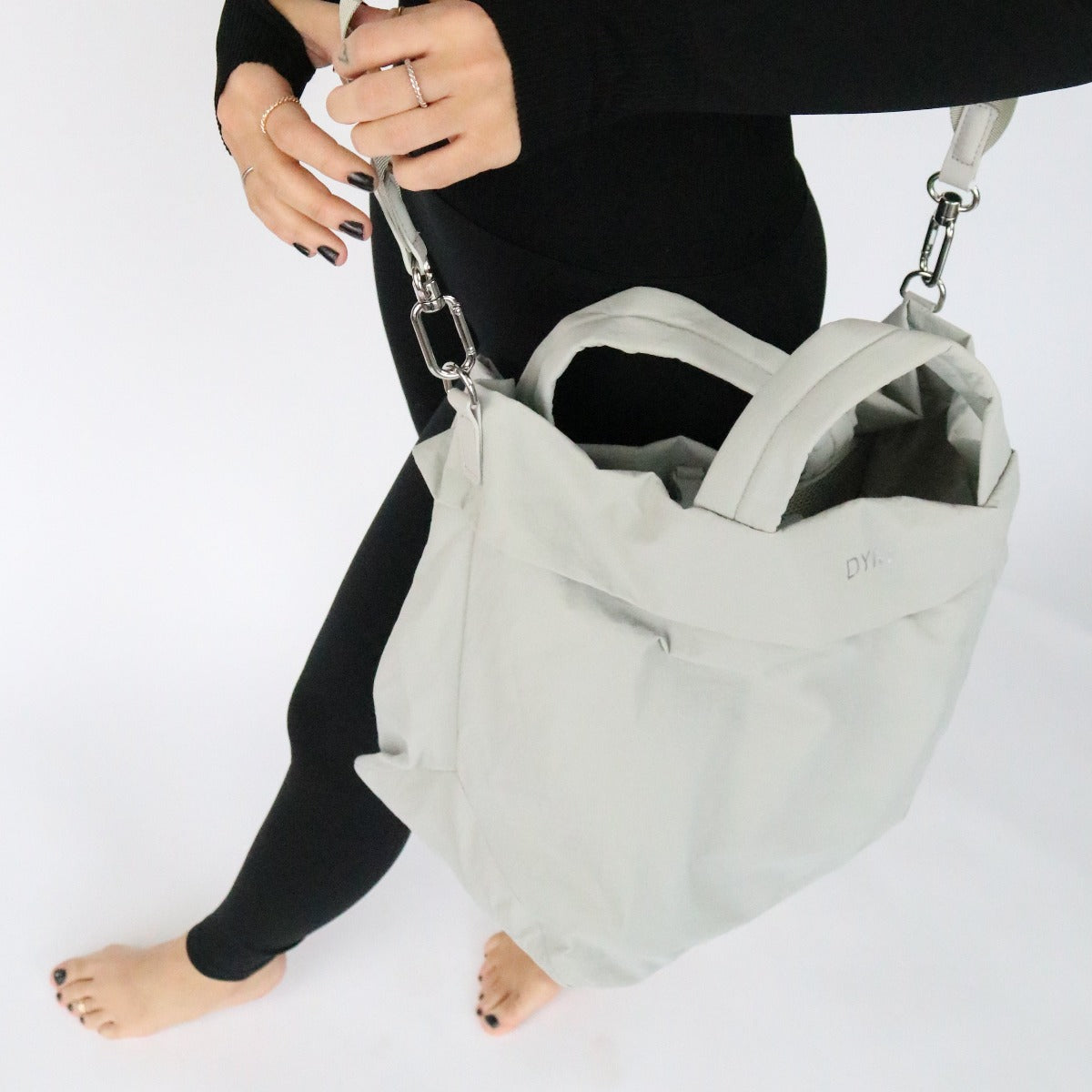A person in black clothes and barefoot holds the DYM Athletics In Motion Bag—a large light gray work-to-gym bag with hand and shoulder straps—at their side, showing only their lower body and ringed, black-polished right hand against a white background.