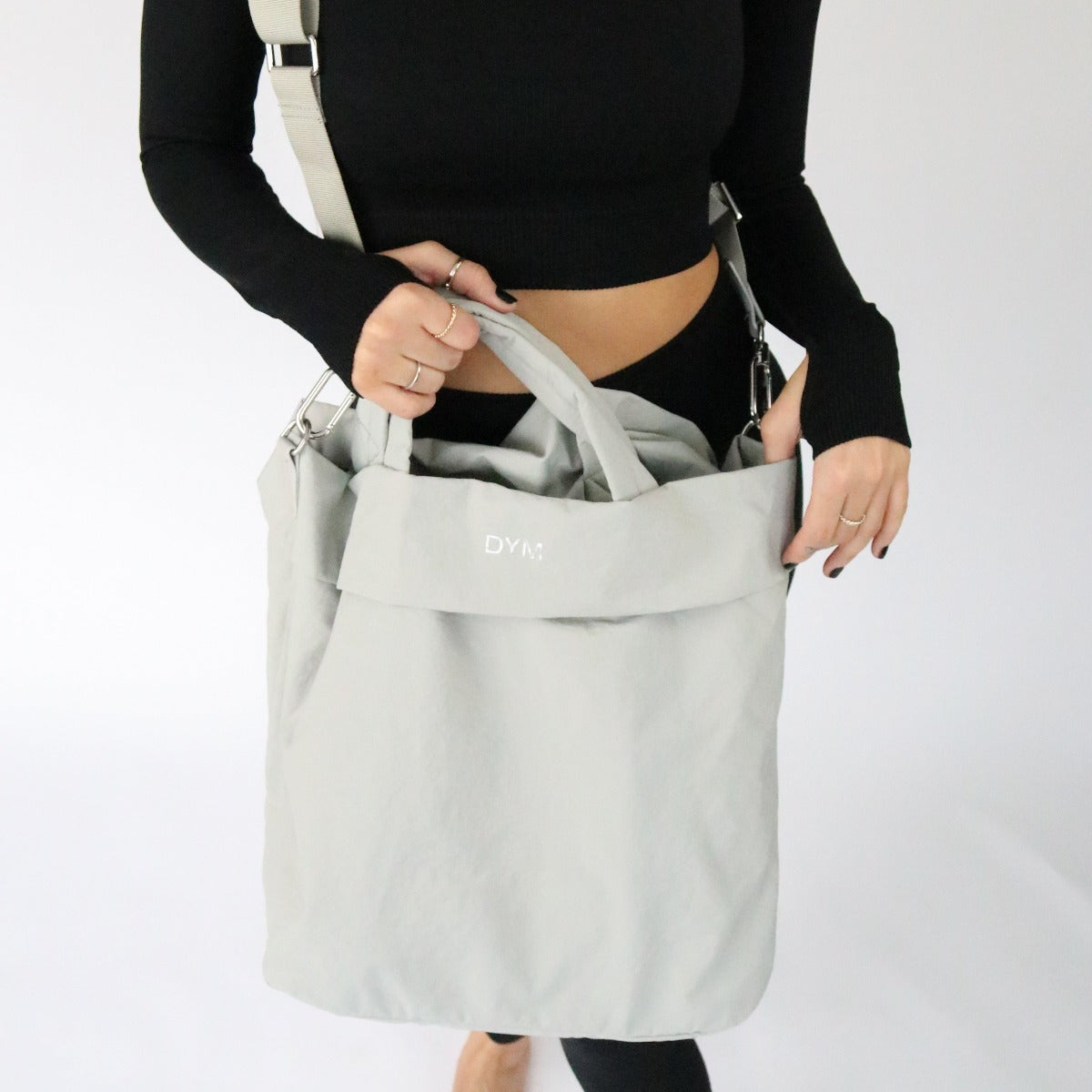 A person with painted nails and rings holds the In Motion Bag by DYM Athletics, a light gray travel bag with silver hardware and DYNE branding on the front. They're dressed in a fitted black long-sleeve top against a plain white background.