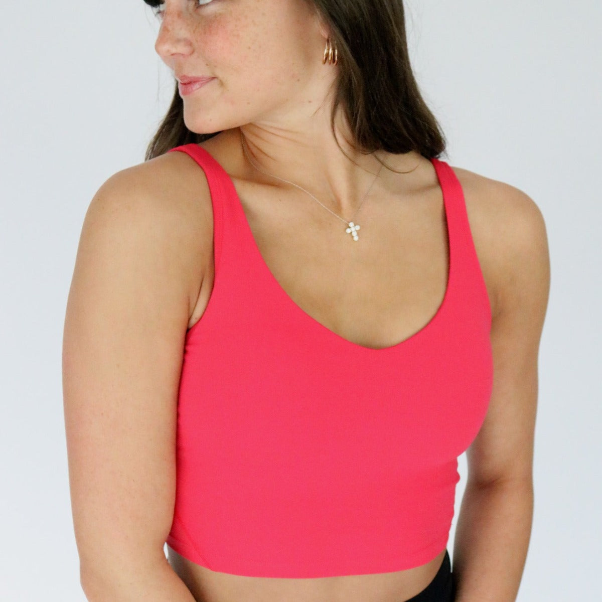 A woman with long brown hair and fair skin wears a bright pink DYM Athletics Flex V-Neck Bra with Scoop Back (medium support, A-D+ cup, SALE) and a gold cross necklace. She looks left and smiles slightly against a plain light background.
