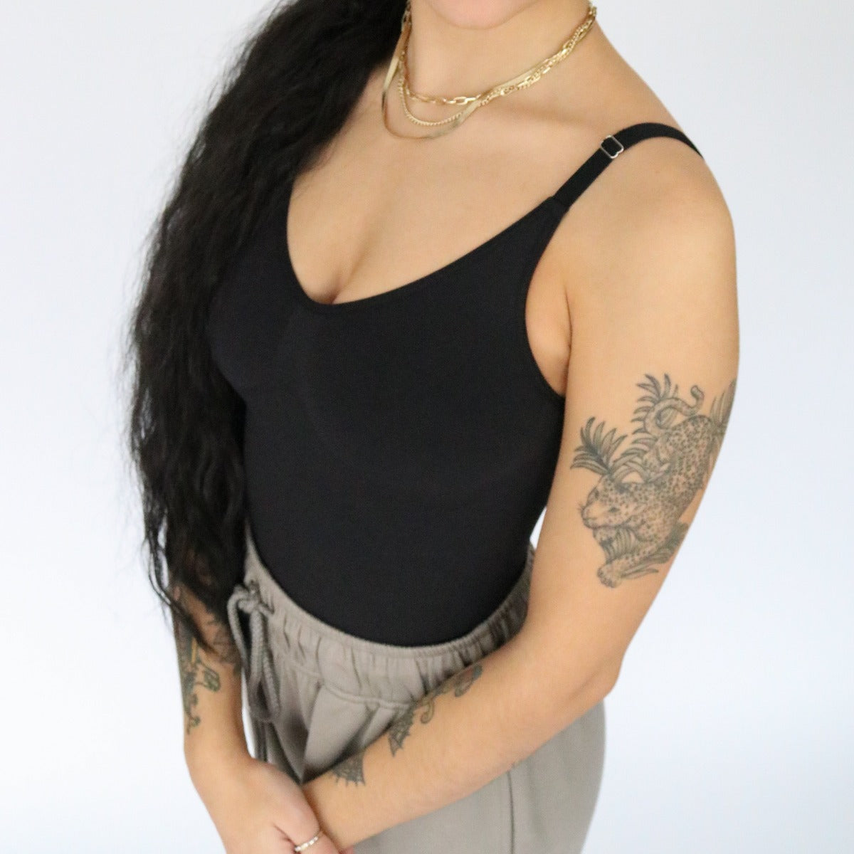 A person with long, dark wavy hair wears the DYM Athletics Shapewear Bodysuit and beige drawstring pants. Tattooed arms, layered gold necklaces, and a light background; photo cropped above the nose.
