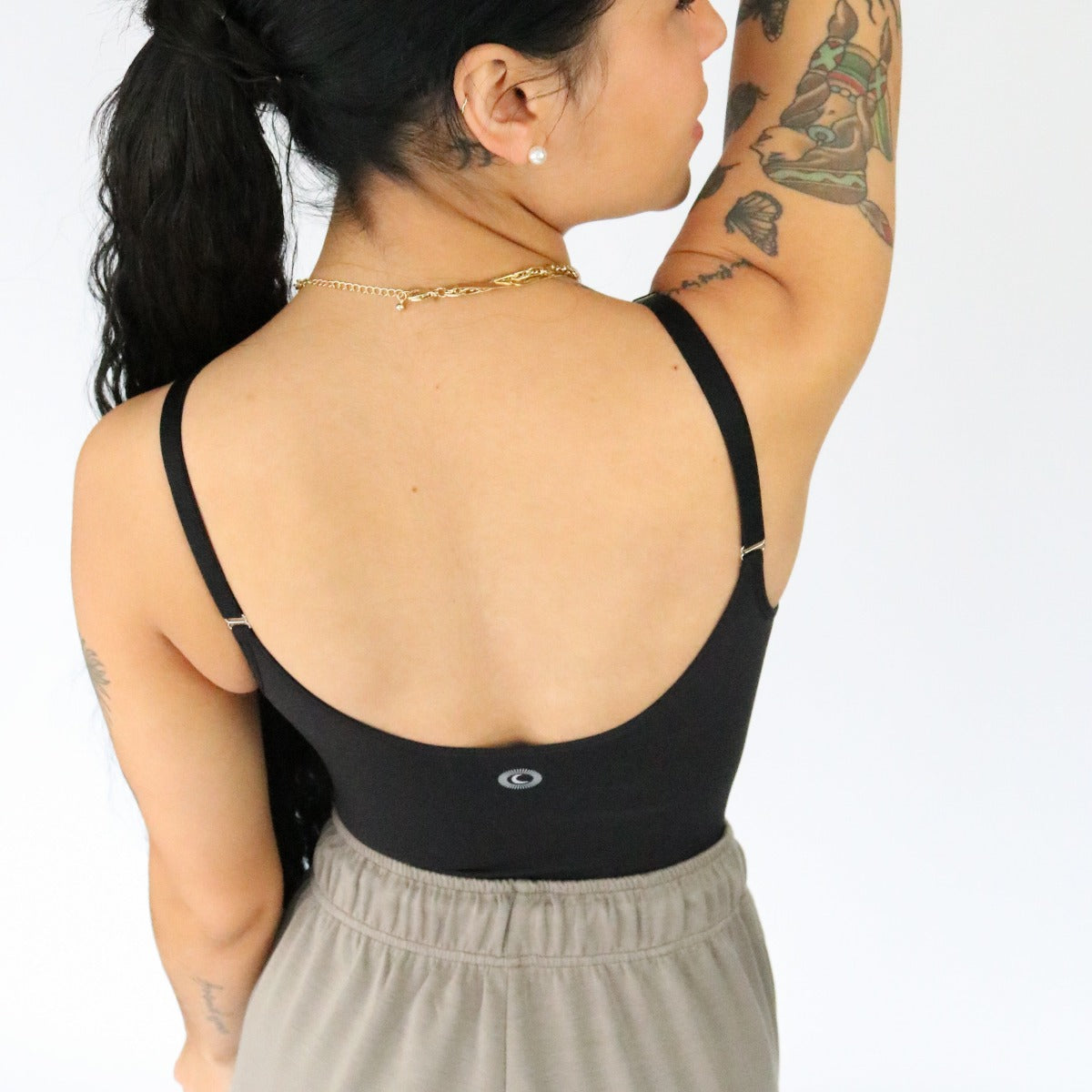 A woman with long dark hair in a ponytail stands with her back to the camera wearing the DYM Athletics Shapewear Bodysuit, showing off colorful tattoos, a gold chain necklace, and small stud earrings.