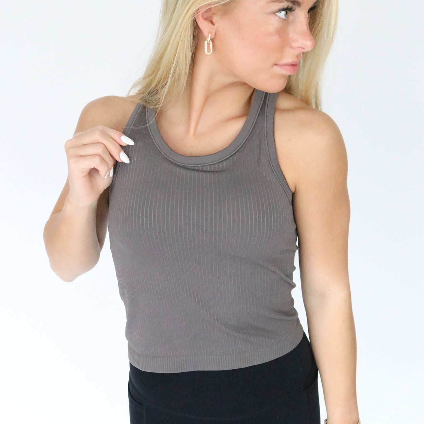 A woman with long blonde hair wears the DYM Athletics Restore Ribbed Comfort Tank w/ Built in Bra and black pants, standing against a white background. She looks right, holds her tank strap, and wears small hoop earrings.