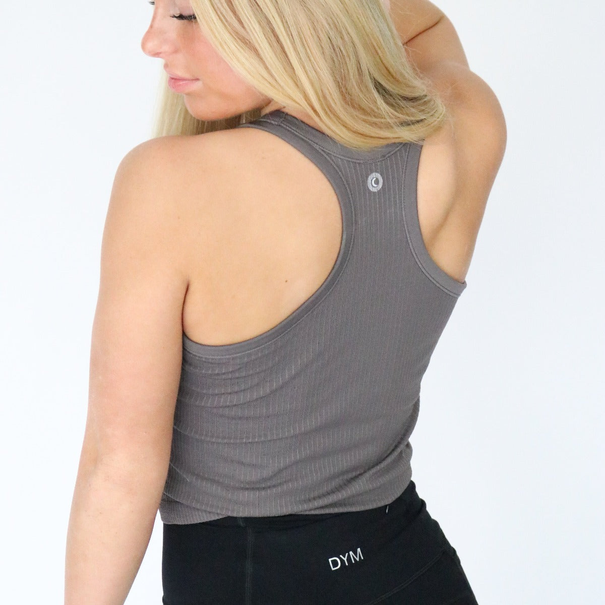 A woman with long blonde hair faces away, wearing DYM Athletics' Restore Ribbed Comfort Tank w/ Built in Bra and high-waisted black leggings with a “DYM” waistband. The ribbed gray top offers a supportive bralette. Background is plain white.
