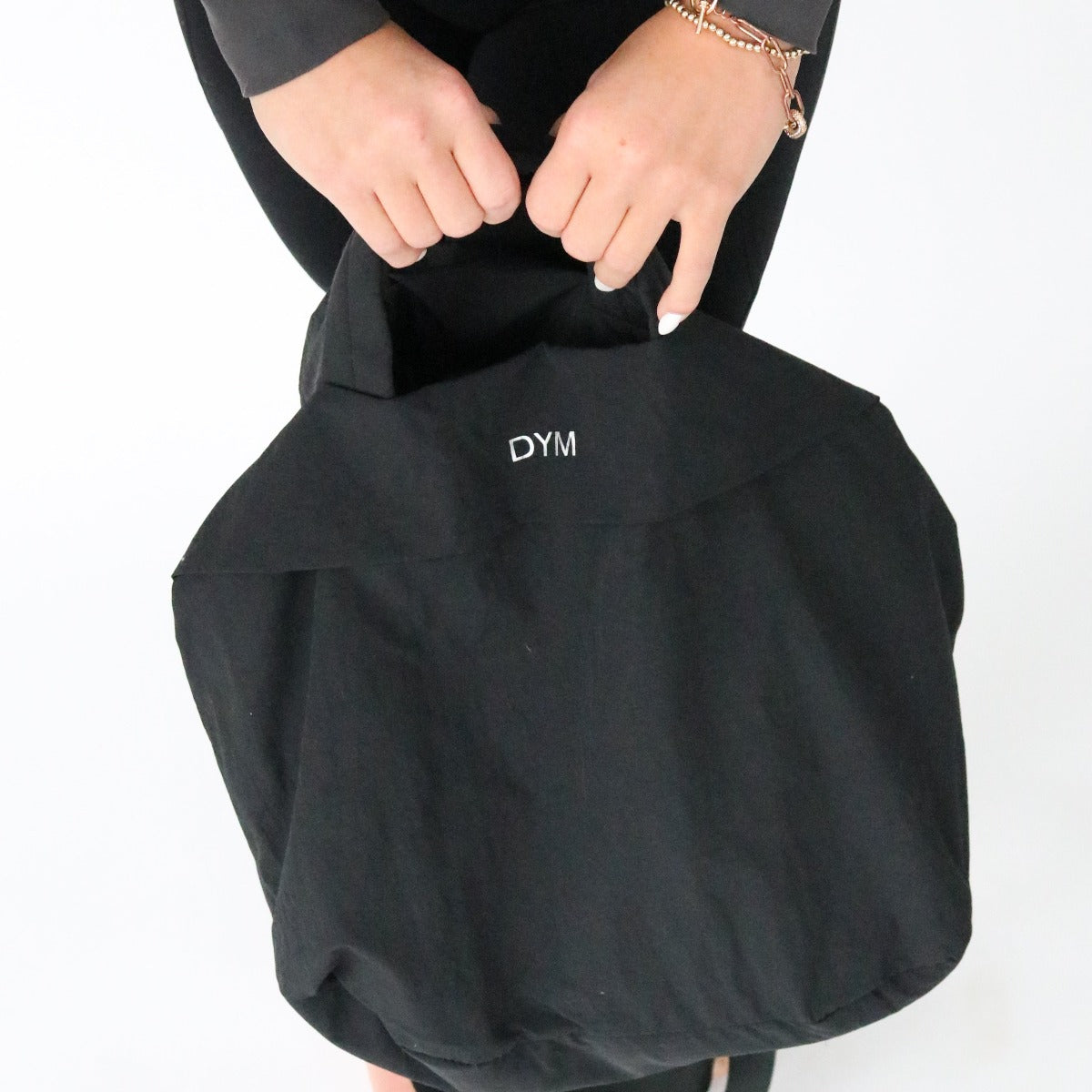 A person with light skin holds the black In Motion Bag by DYM Athletics, featuring white "DYM" embroidery, in both hands. They're dressed in black with a rose gold bracelet, standing before a plain white background.