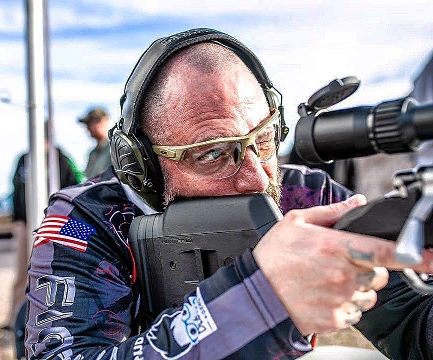 A man in camouflage and earmuffs aims a rifle at an outdoor range, wearing Epoch Eyewear Grunt Tactical Sport Sunglasses for eye protection.