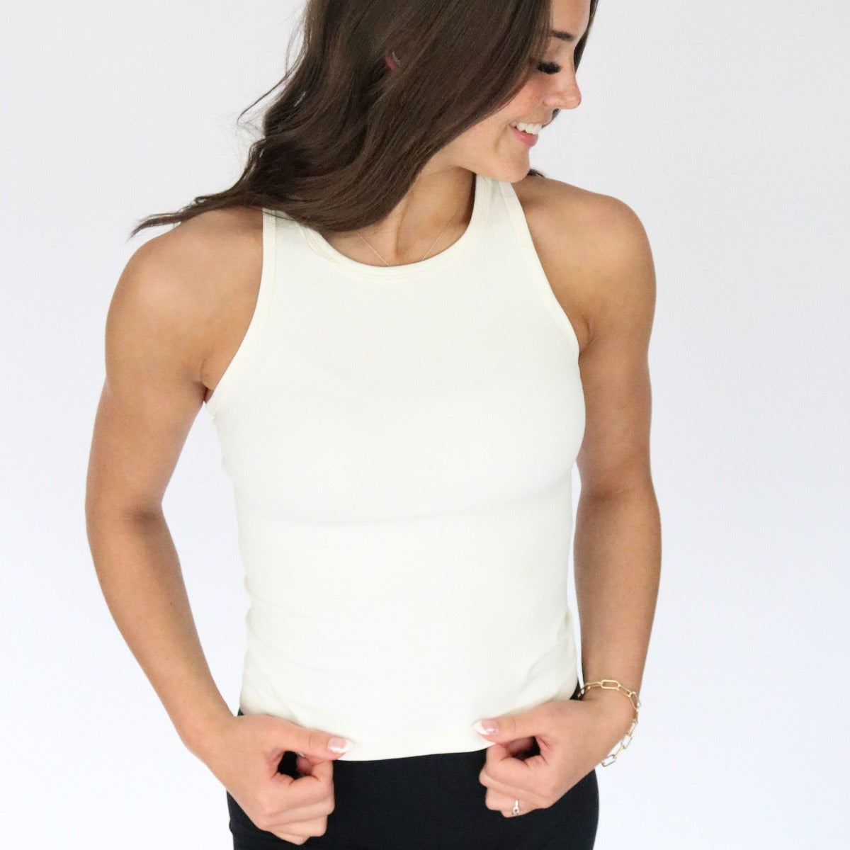 A woman with long brown hair smiles as she looks down, holding the sides of her Go-To Waist-Length Racerback Tank Top by DYM Athletics. She pairs it with black pants and a bracelet on her left wrist against a plain white background.