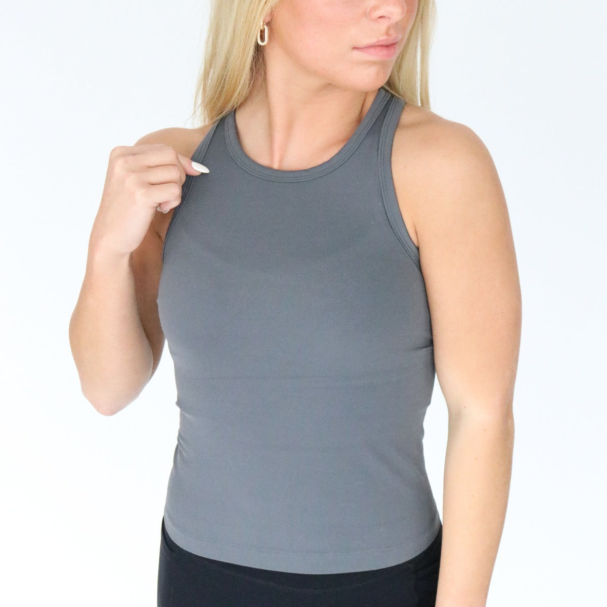 A woman with long blonde hair wears the DYM Athletics Go-To Waist-Length Racerback Tank Top with fitted gray and black bottoms. She looks to her right, holding the left strap of her top against a plain white background.