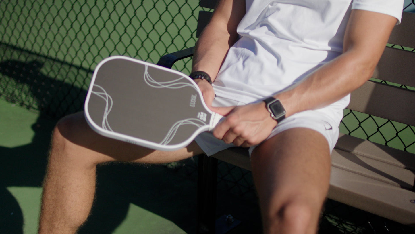 A person in a white shirt and shorts sits on a bench, holding the LUXE Pickleball Finesse carbon fiber paddle on their lap. Their face isn’t visible. They wear a smartwatch and fitness band; a chain-link fence is behind them.