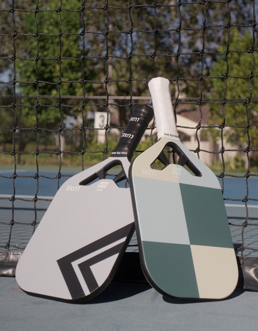 Two LUXE Pickleball Draft paddles, including a carbon fiber model, lean against an outdoor net. One is light gray with black stripes; the other sports green, cream, and gray blocks. Trees, a fence, and court surface appear in the blurred background.