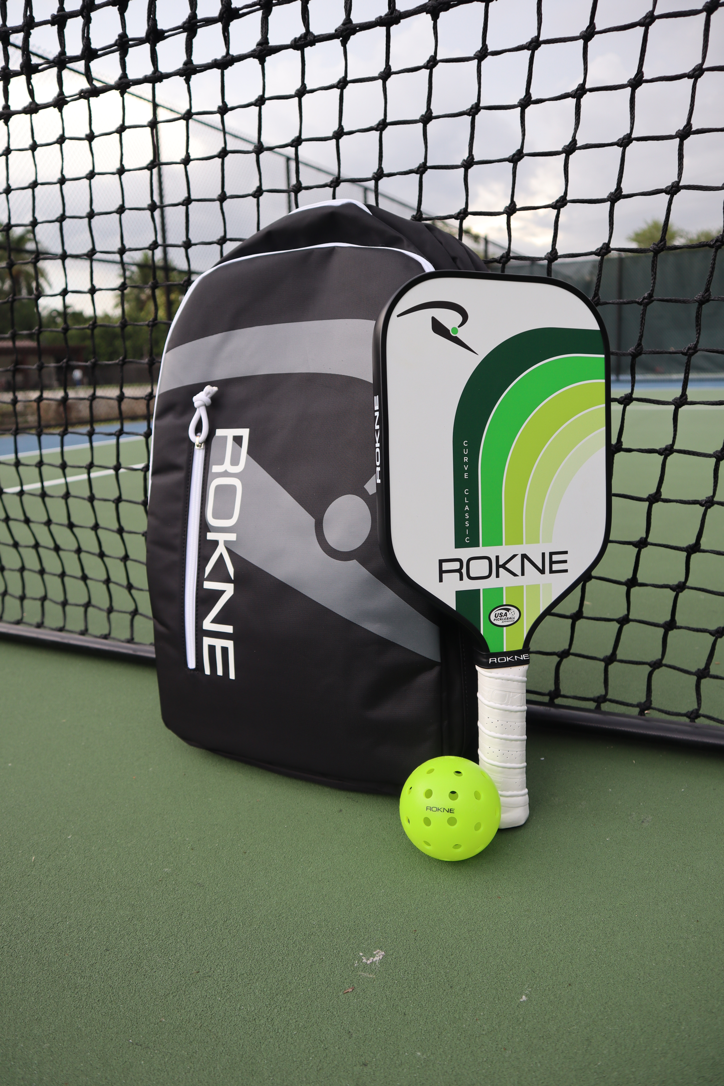 A black ROKNE BACKPACK, a green and white ROKNE paddle, and a neon yellow pickleball rest on the green court by the net, with tennis fencing and cloudy skies behind—ideal for carrying your pickleball gear.