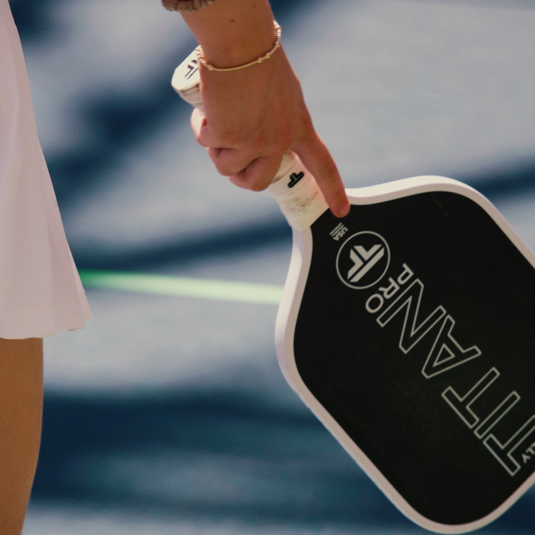 Close-up of a person gripping a Rally Titan Pickleball Titan Power 14MM paddle with T700 Titan Carbon Friction Surface, wearing a white skirt and gold bracelet. Blue court surface and sharp green line appear in the background.