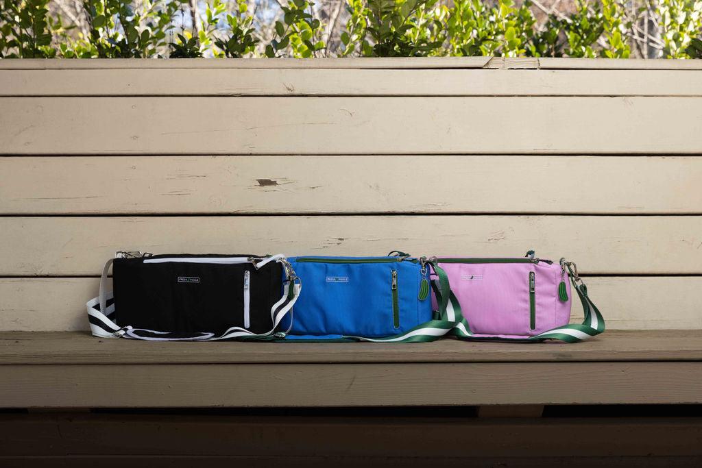 Three Fresh Pickle Insulated Cooler Bags—black, blue, and pink—sit on a wooden bench. Each bag features zippered pockets and detachable straps, keeping court essentials organized. Sunlight and green foliage brighten the scene.