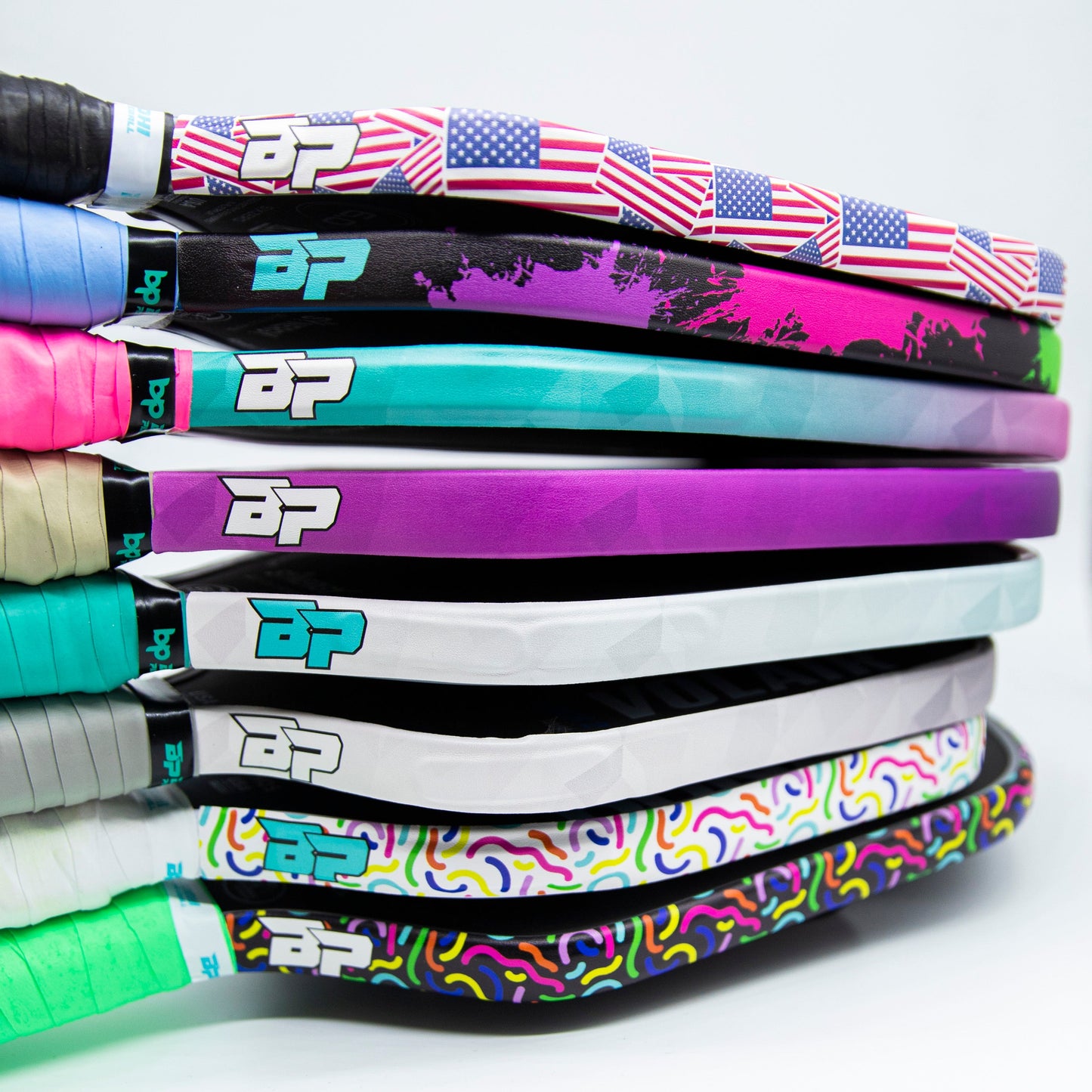 A close-up of eight stacked tennis rackets with colorful Bodhi Performance Premium Edge Tape - PATTERNS, featuring American flag, gradient, camo, and abstract designs plus the BP logo near the handle for a vibrant look.