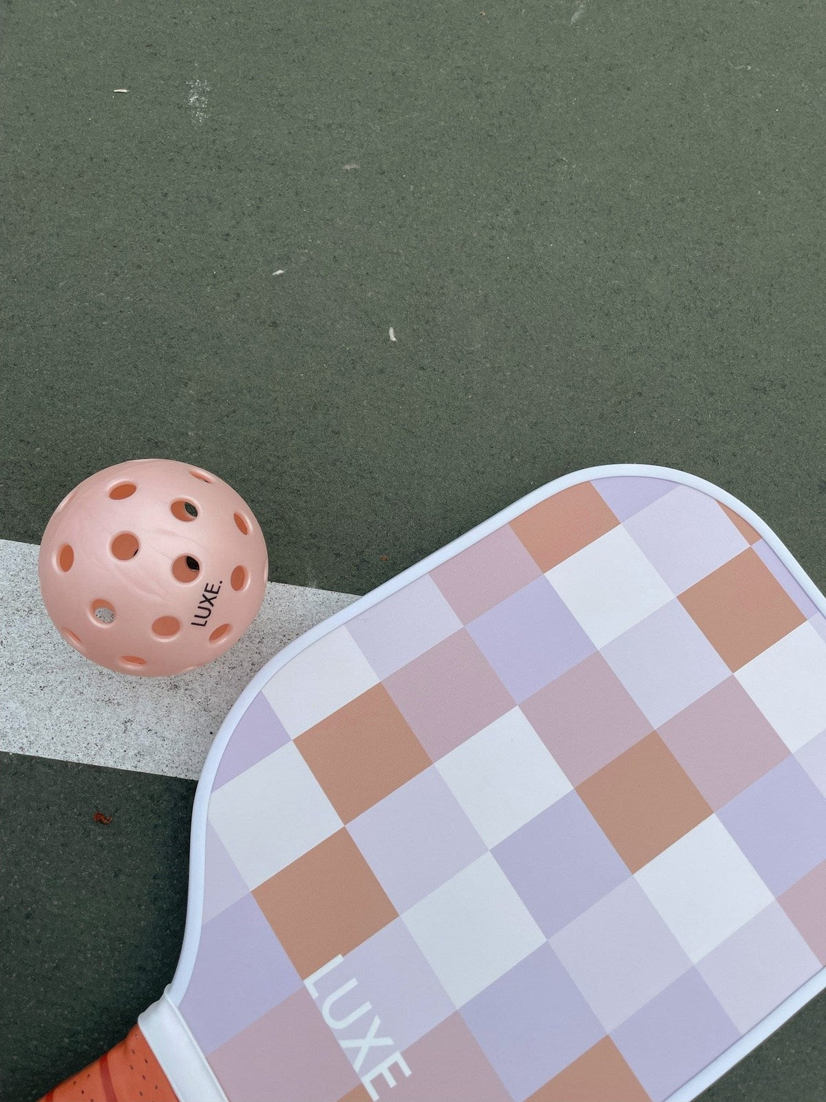 A pink pickleball with holes and LUXE Pickleball branding sits beside the Checker paddle—by LUXE Pickleball—which features pastel peach, pink, and lavender squares plus a comfy grip band on a green outdoor court.