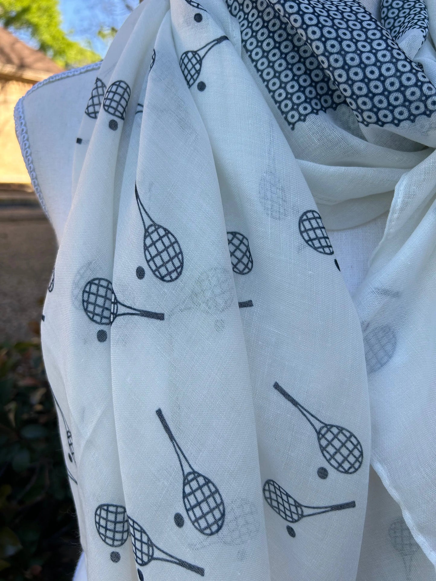 The Racquet Time Sun Scarf/Wrap by Runway Athletics, shown draped on a mannequin, features black racket motifs over white with a bold geometric circle border—sunlight highlighting this stylish sun protection accessory against blurred greenery.