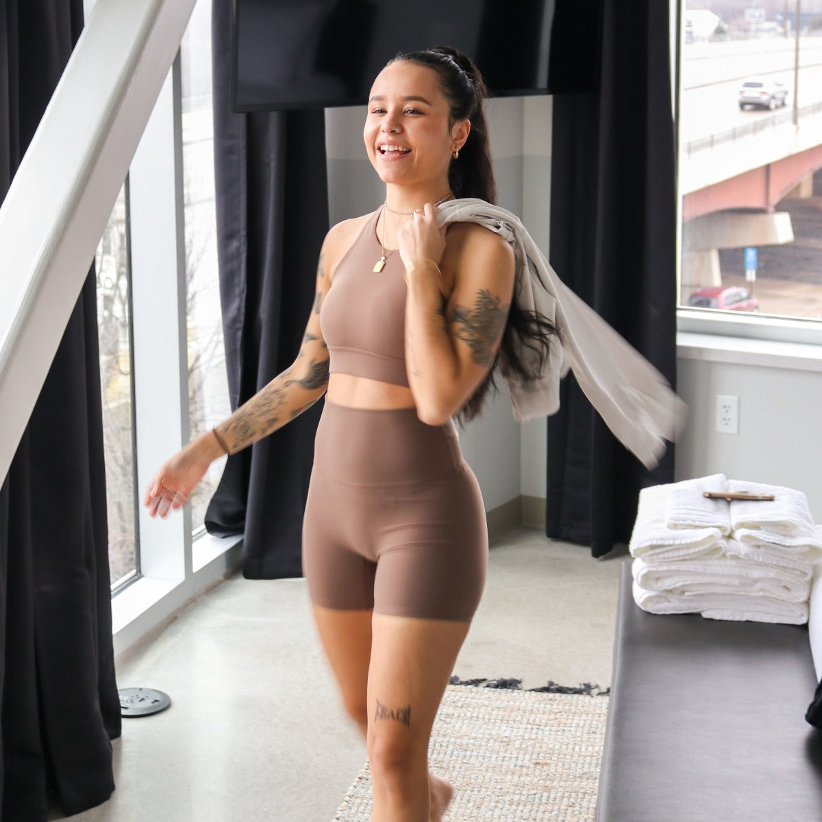 A woman with long dark hair and tattoos smiles as she walks indoors, wearing DYM Athletics' AIRE Ultra-Soft High Waist Biker Shorts | 5" in taupe with a matching sports bra, holding a towel over her shoulder. Large windows show an urban street outside.