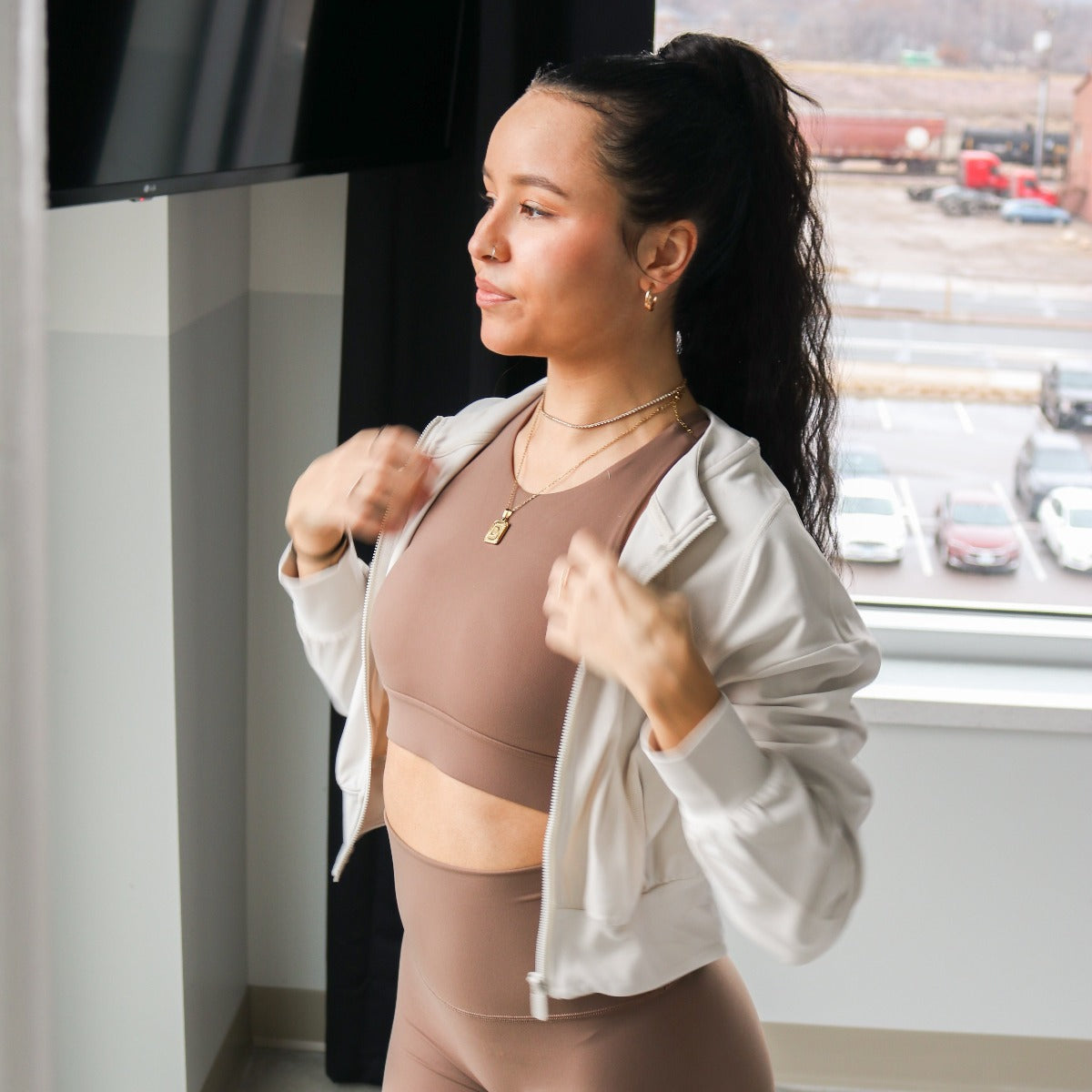 A woman with long dark hair in a ponytail stands by a window, wearing the DYM Athletics AIRE Ultra-Soft Dynamic Adjustable Bra (high support, A-D+ cup) and leggings with a light zip-up jacket. She adjusts her jacket, looking sideways. Cars and a railway are outside.