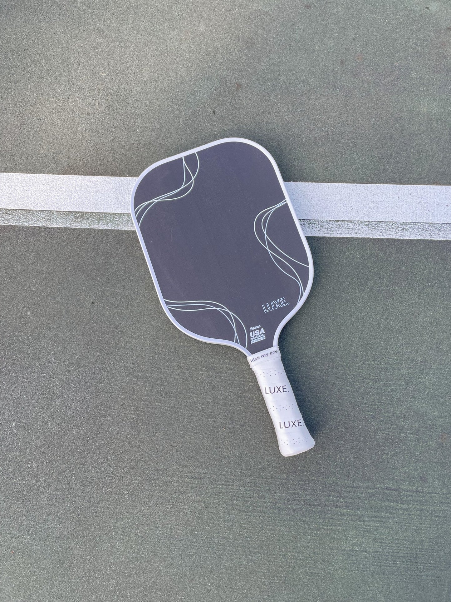 A black carbon fiber LUXE Pickleball Finesse paddle with white abstract lines and LUXE branding lies on a green court, partly over a white line. The handle features a white “LUXE” grip tape for enhanced control.