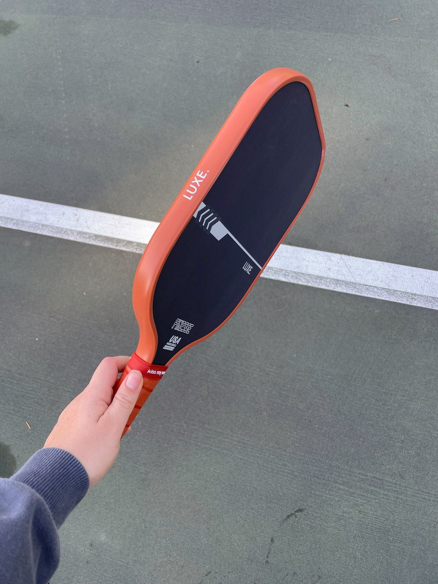A person in a gray sleeve holds a black and red carbon fiber LUXE Pickleball Finesse paddle over a green outdoor court, with a white boundary line visible in the background.