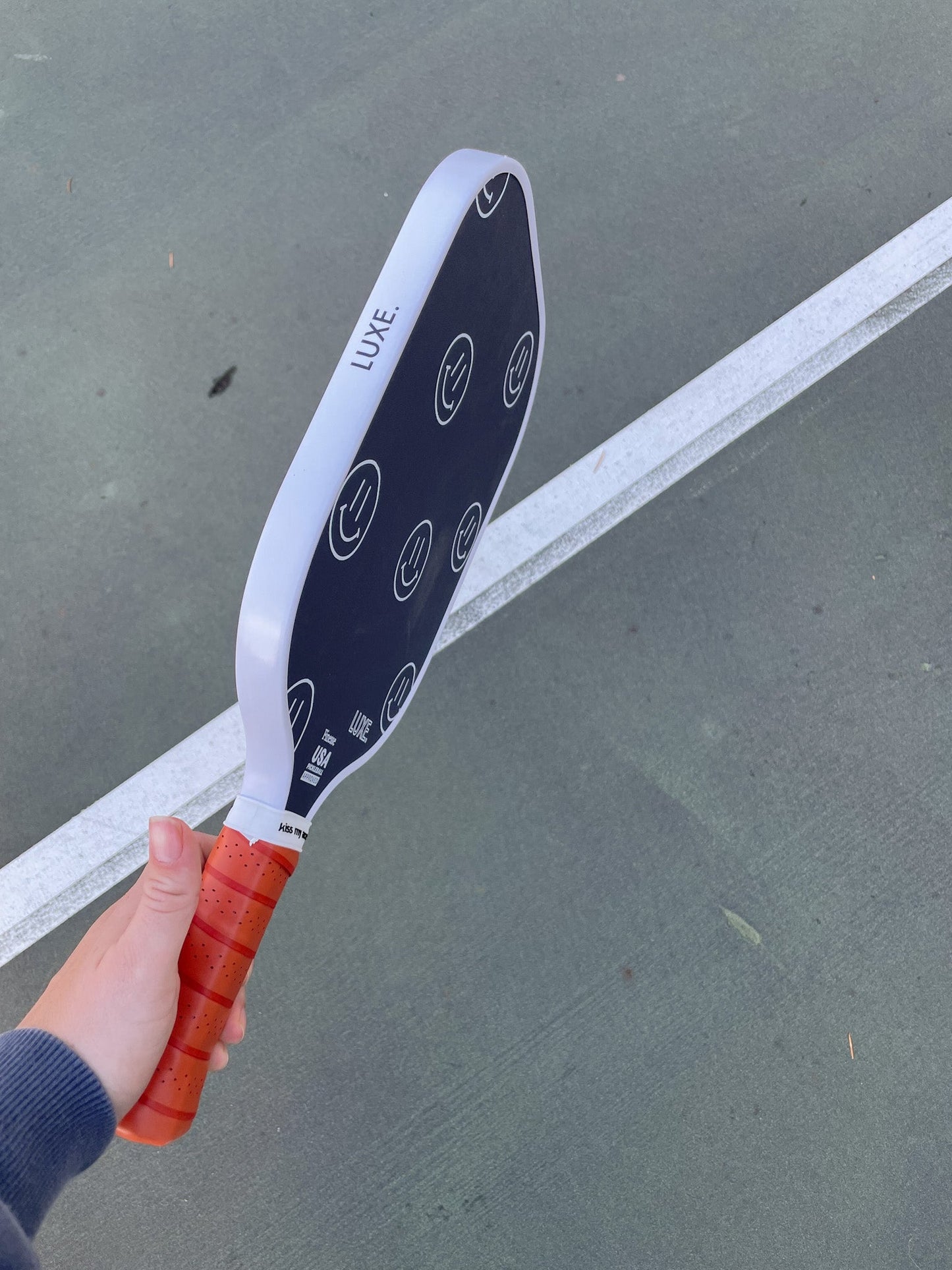 A hand holds the LUXE Pickleball Finesse paddle, featuring a black carbon fiber face with white smiley faces, an orange grip, and “LUXE” on the edge. A green court with white lines highlights its premium design for unmatched control.