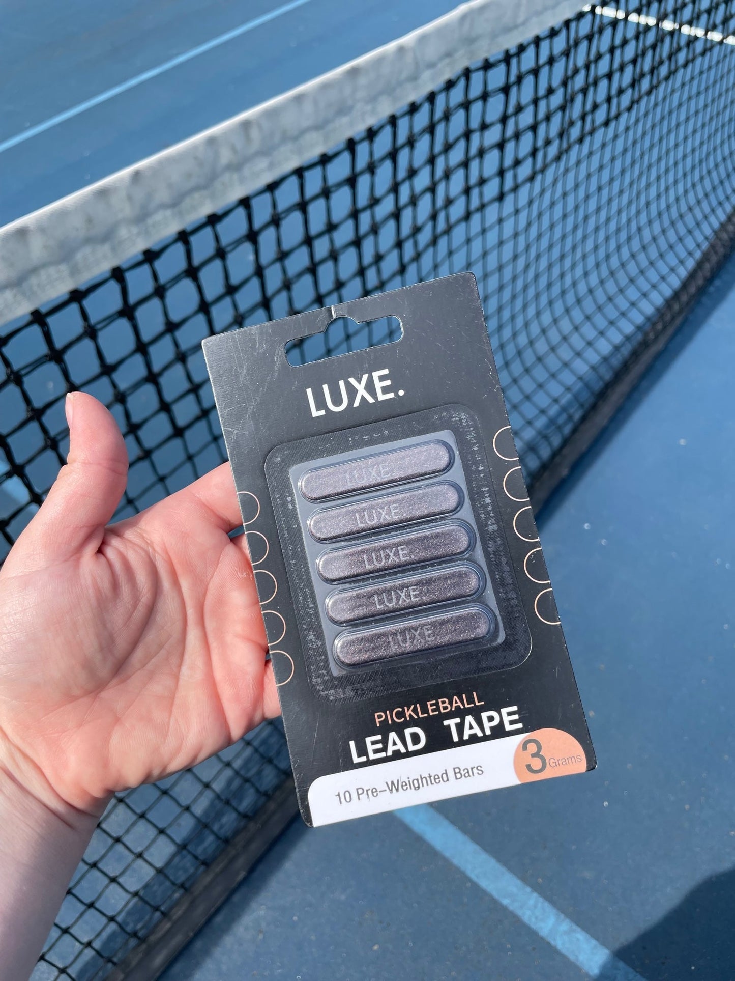 A hand holds a LUXE Pickleball Lead Tape package with 10 pre-weighted 5g strips in front of an outdoor court’s black net. The black pack has metallic silver lettering and orange accents, clearly showing the contents.