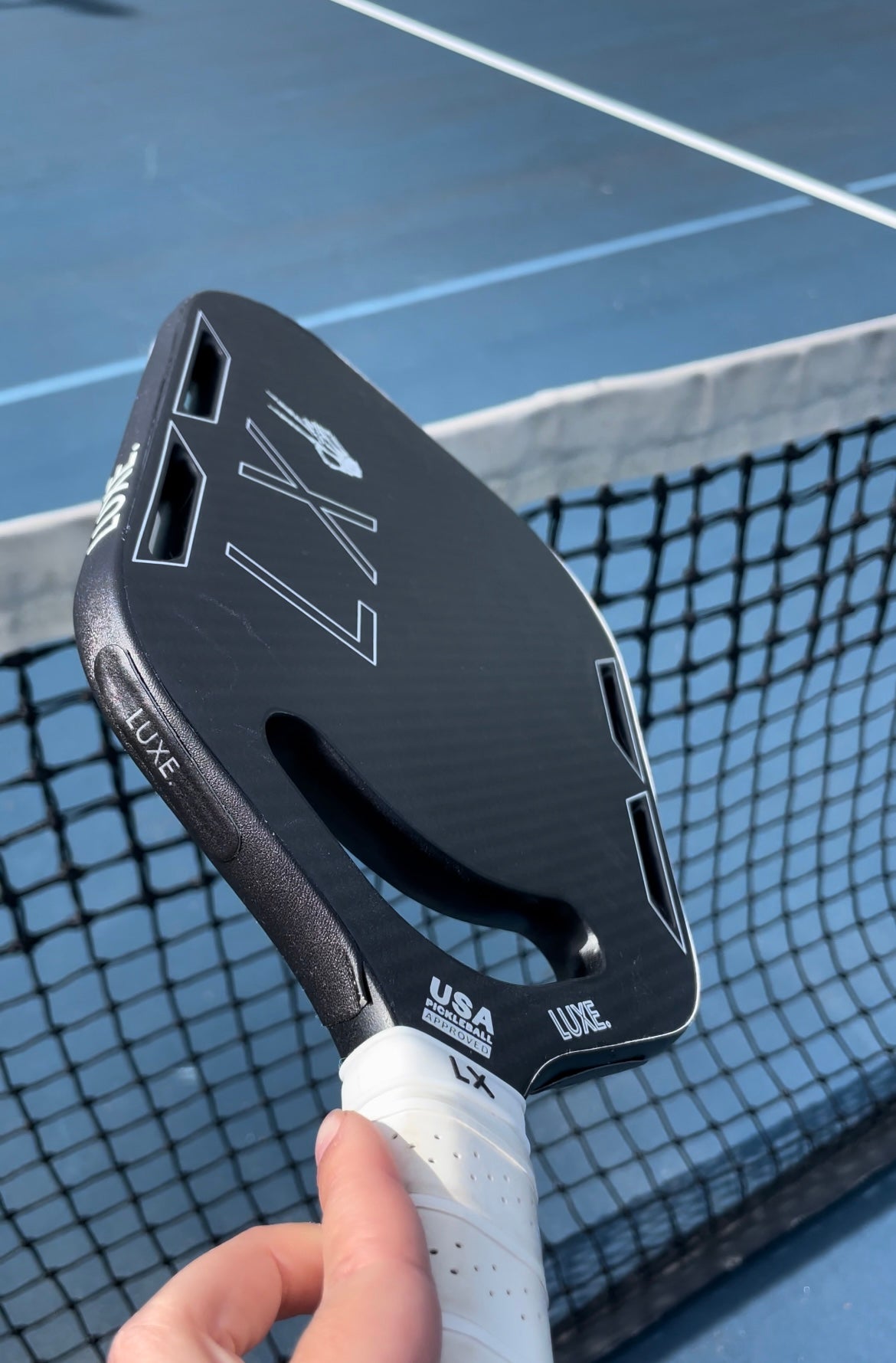 A black carbon fiber LUXE Pickleball paddle with a white grip and 5g Lead Tape added to the edge is held in front of a net on a blue court. “USA Pickleball Approved” is shown on the paddle’s edge.