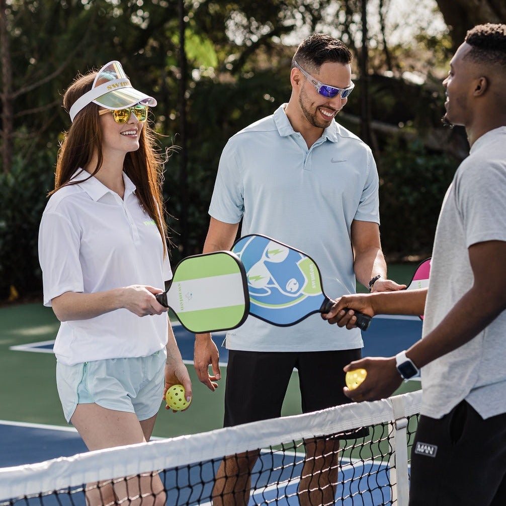 Three adults stand on a pickleball court by the net, smiling. One woman in a visor and sunglasses holds a Swinton Pickleball Hero Blue Dog Paddle, while two men with graphite paddles chat nearby. Trees and greenery fill the background.
