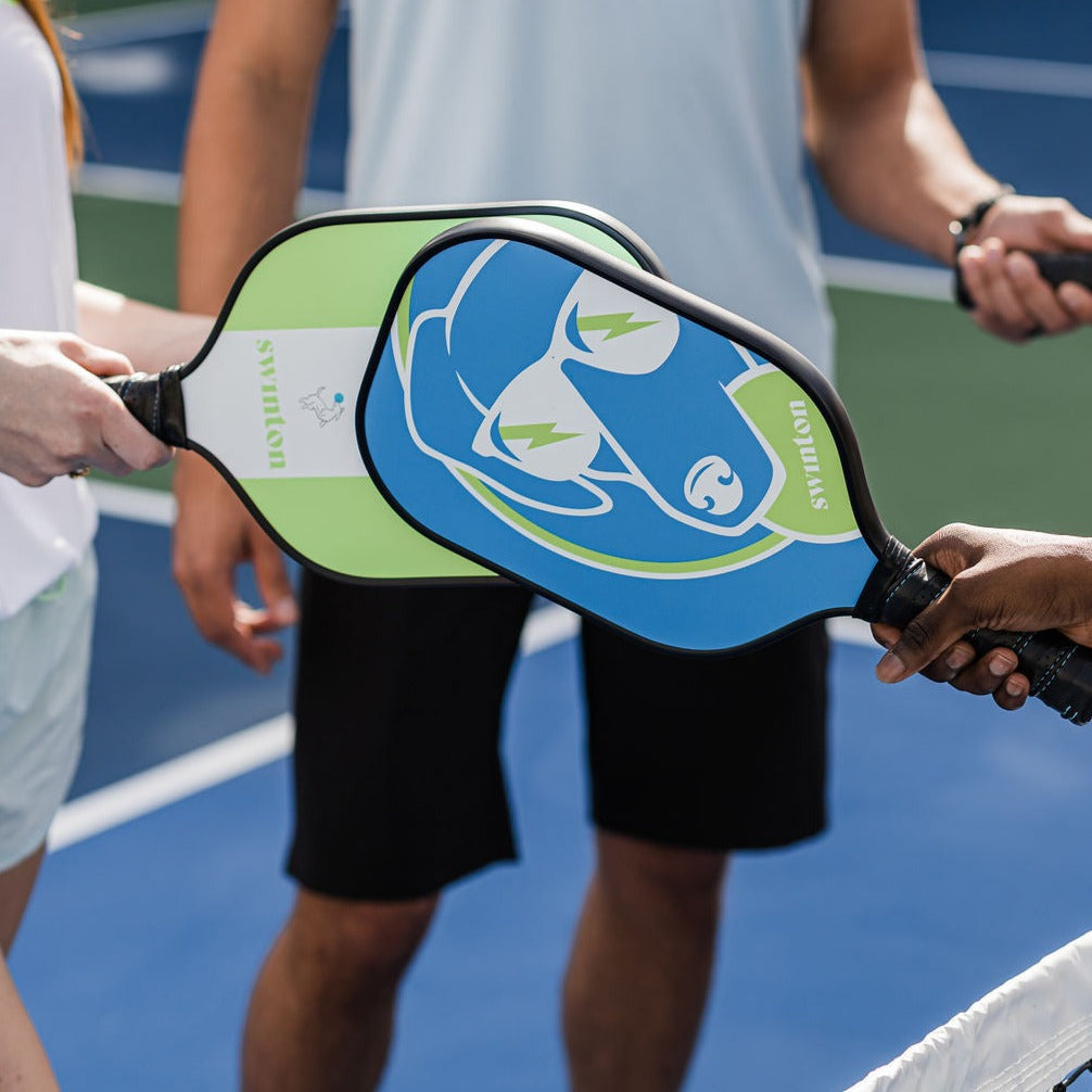 Three people stand on a blue pickleball court, joining paddles at the center. A Swinton Pickleball Hero Blue Dog Paddle is visible alongside a graphite paddle; only arms and sportswear appear, faces out of frame.