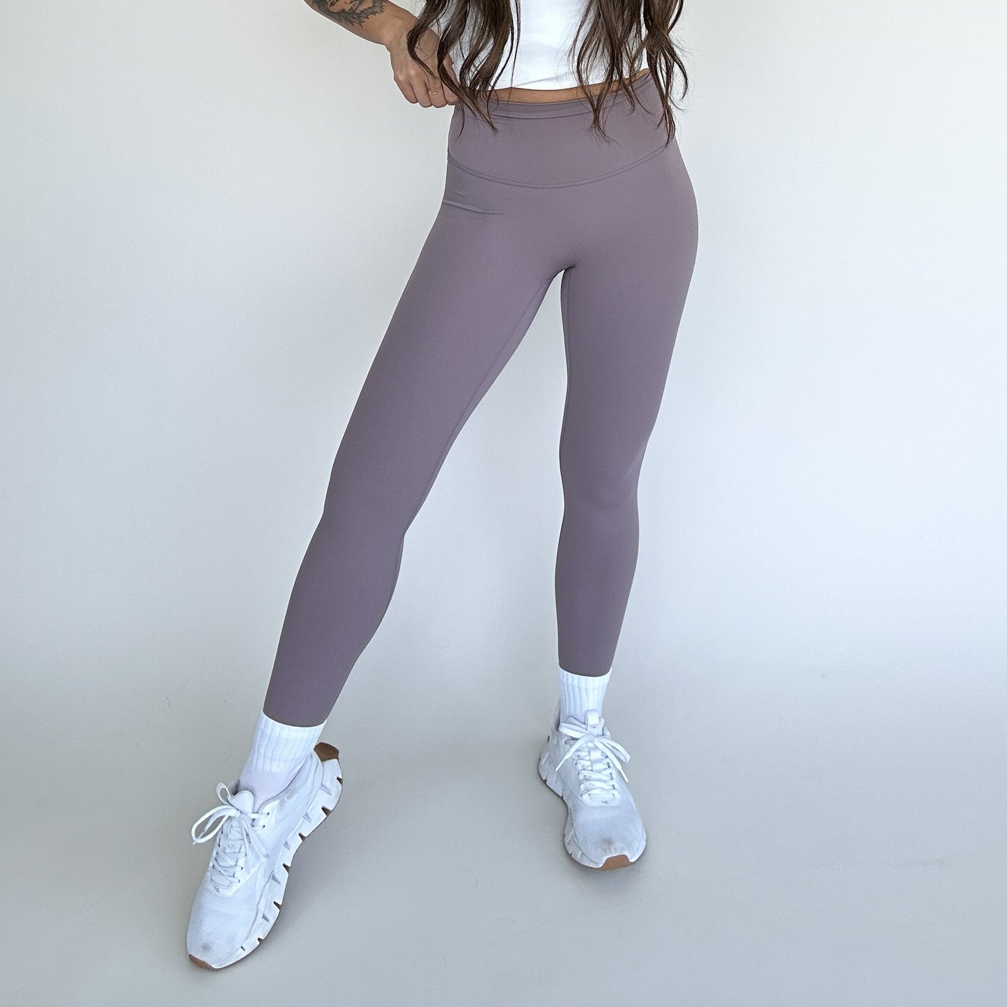 A woman poses against a plain light background in DYM Athletics Flex & Flow mauve high-waisted leggings, a white crop top, white athletic socks, and chunky sneakers. Her long hair is down and one hand rests on her waist for this sporty look.