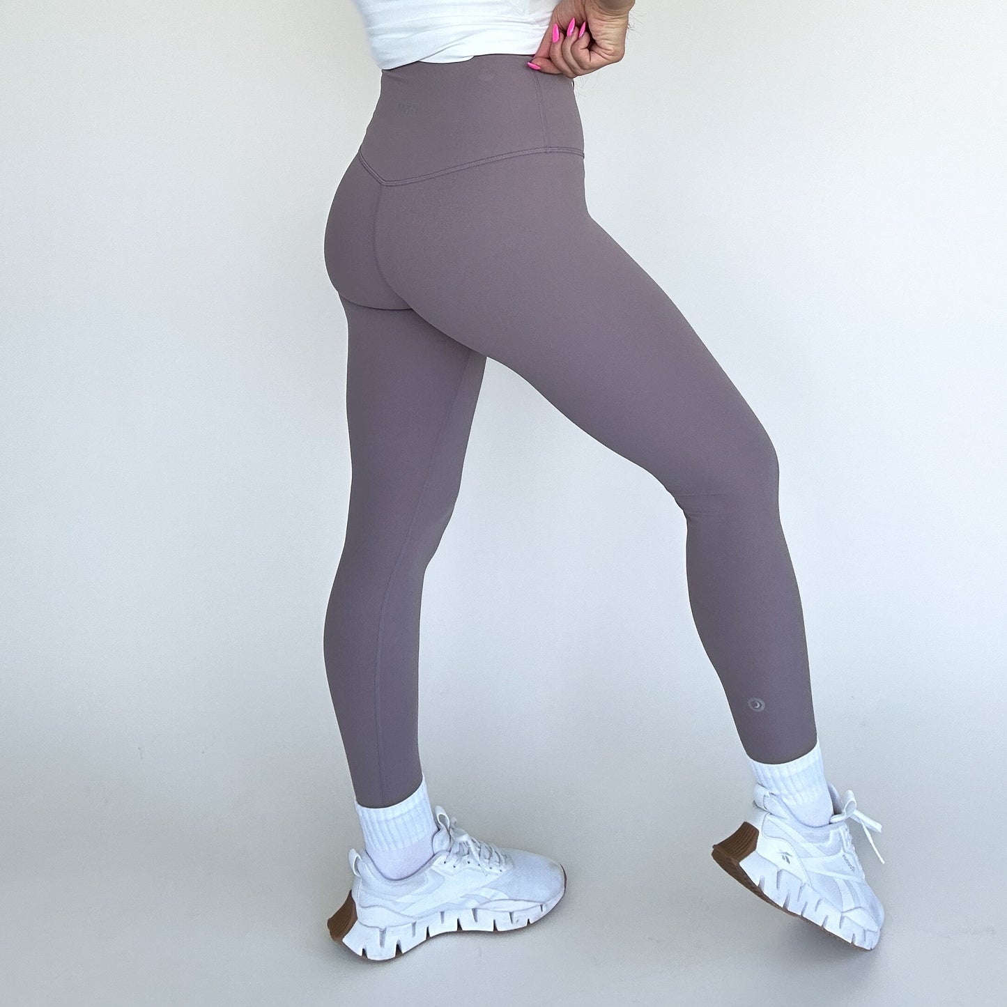 A person stands sideways against a plain white background, wearing DYM Athletics Flex & Flow Legging in mauve, paired with a white cropped top, white socks, and athletic shoes. One hand with painted pink nails rests on their hip.