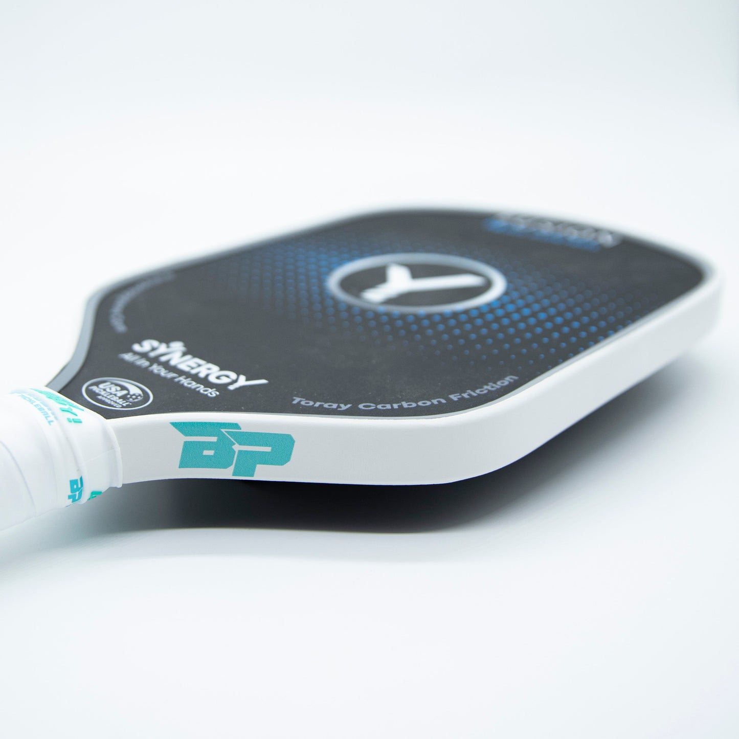 Close-up of a black and blue Bodhi Performance paddle with Premium Edge Tape - SOLIDS for extra protection. The handle has a white grip with teal branding, and the face displays a circular pattern and "SYNERGY." Toray Carbon Fiber visible on edge.