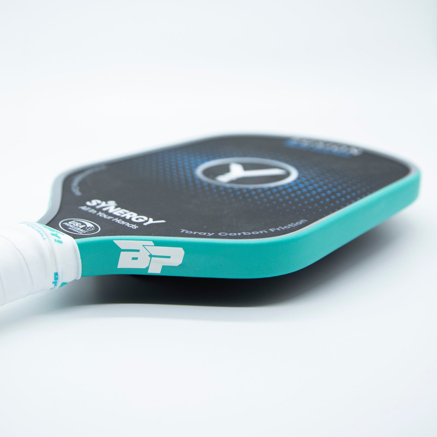 A close-up photo of a Bodhi Performance pickleball paddle with a black and teal face, white grip, and SOLIDS Premium Edge Tape protecting the edge as it lies flat on a white surface, head slightly angled away.